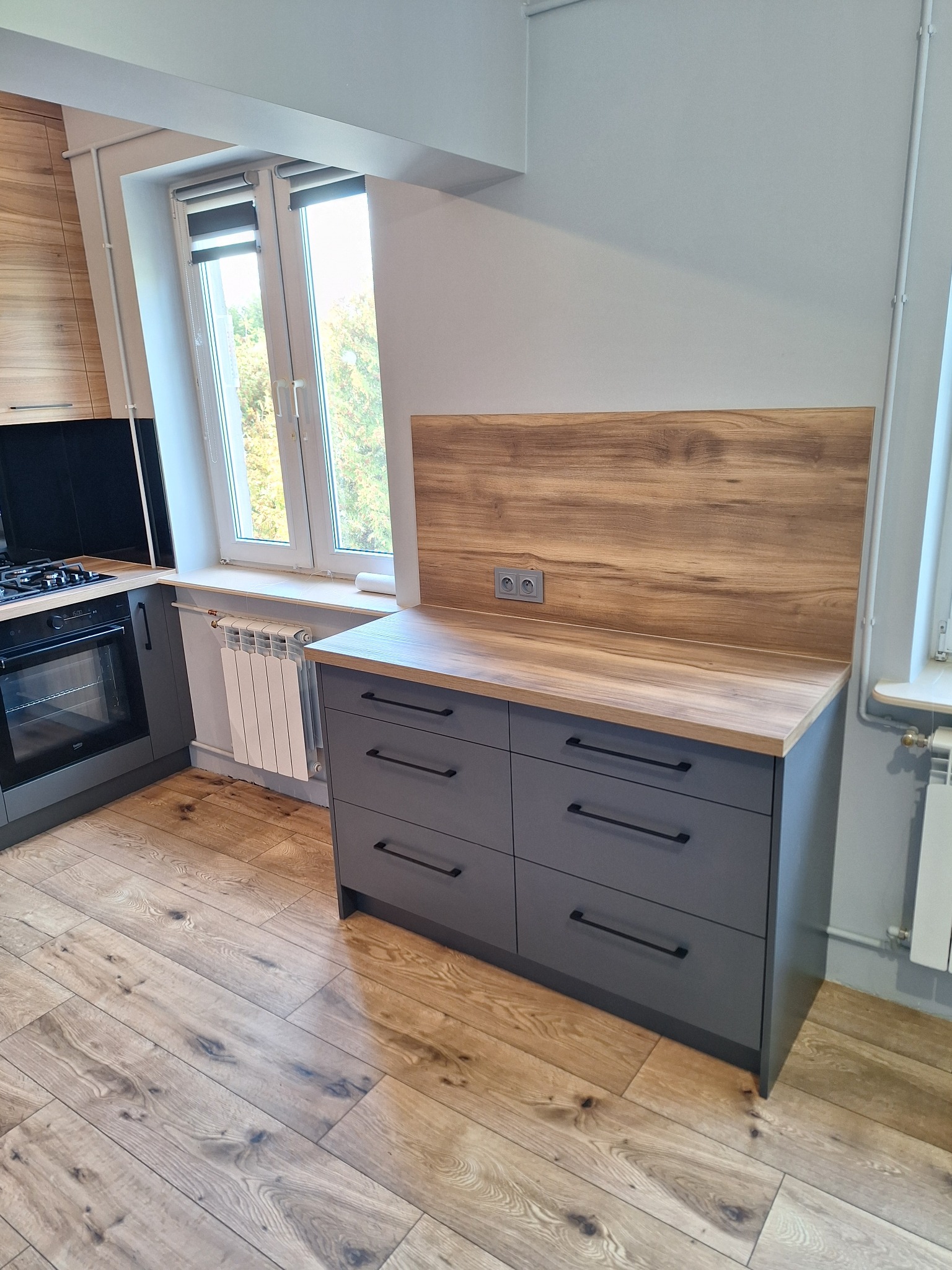 Szara komoda z sześcioma szufladami i drewnianym blatem w kuchni obok okna i grzejnika. Ustawiona na drewnianej podłodze.
