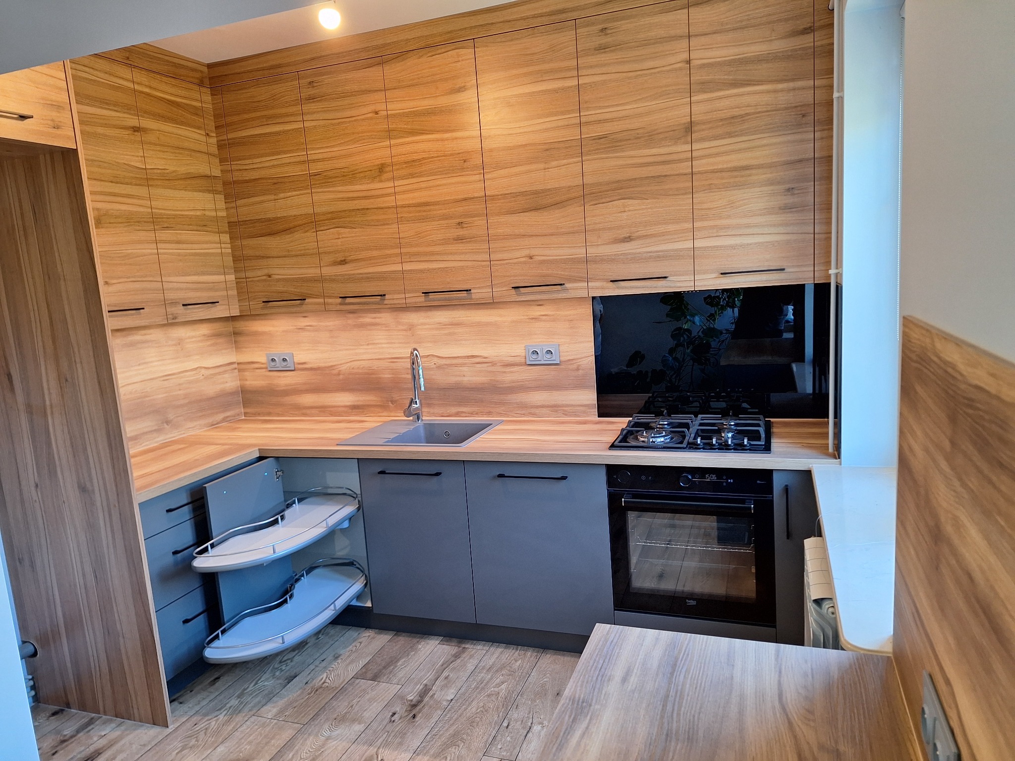 Nowoczesna kuchnia z drewnianymi szafkami górnymi i szarymi dolnymi. Widoczny wysuwany system półek cargo i płyta grzewcza.