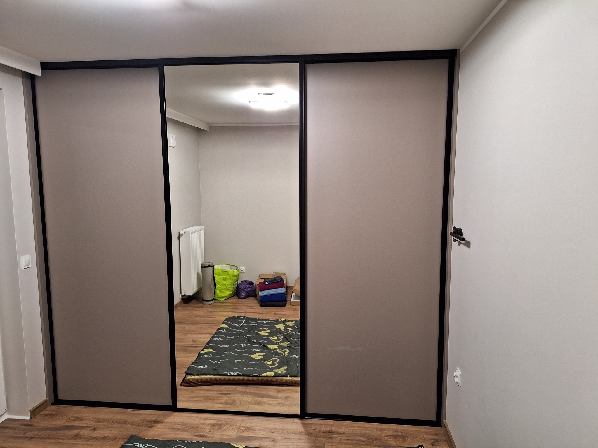 Szafa wnękowa z przesuwnymi drzwiami w odcieniach beżu i czerni, z lustrem na środkowych drzwiach, odbijającym wnętrze pokoju i elementy wyposażenia.