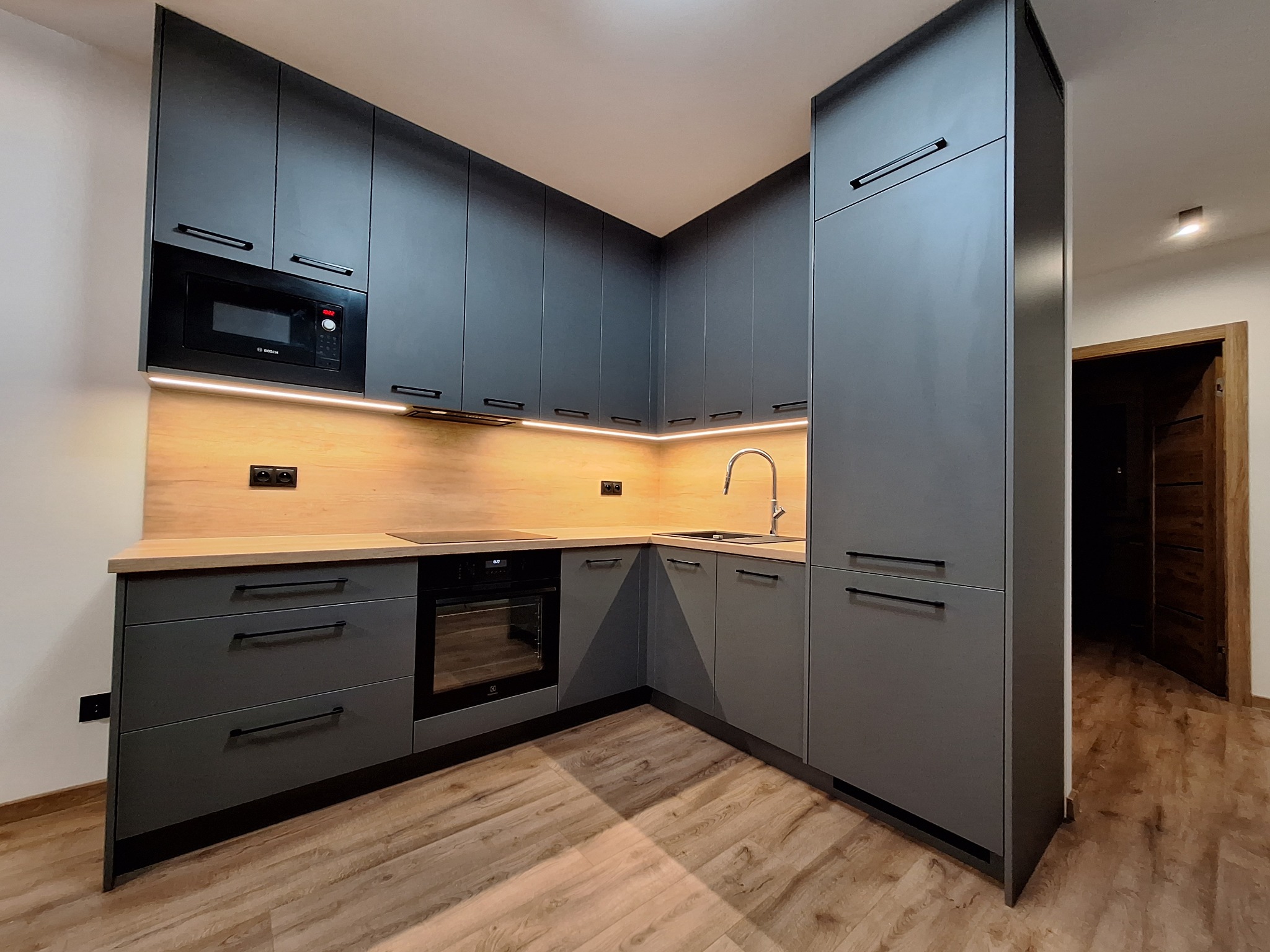 Nowoczesna kuchnia na wymiar w odcieniach szarości z drewnianym blatem i oświetleniem LED, widok z perspektywy podłogi, z widocznym fragmentem drewnianej podłogi.