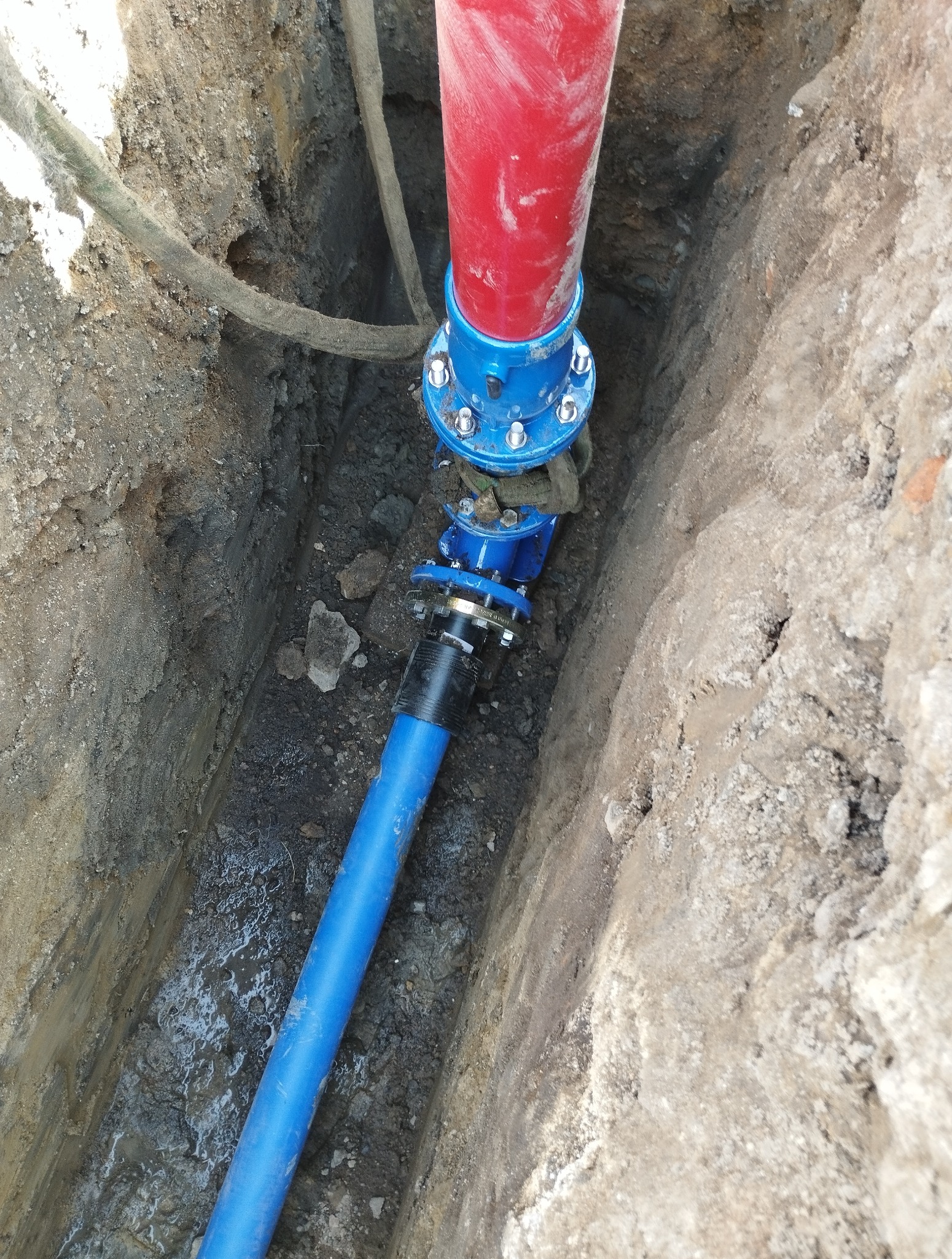 Czerwona i niebieska rura połączona złączką w wykopie. Widoczne ślady świeżych prac ziemnych i wilgoć. Element instalacji wodociągowej lub kanalizacyjnej.