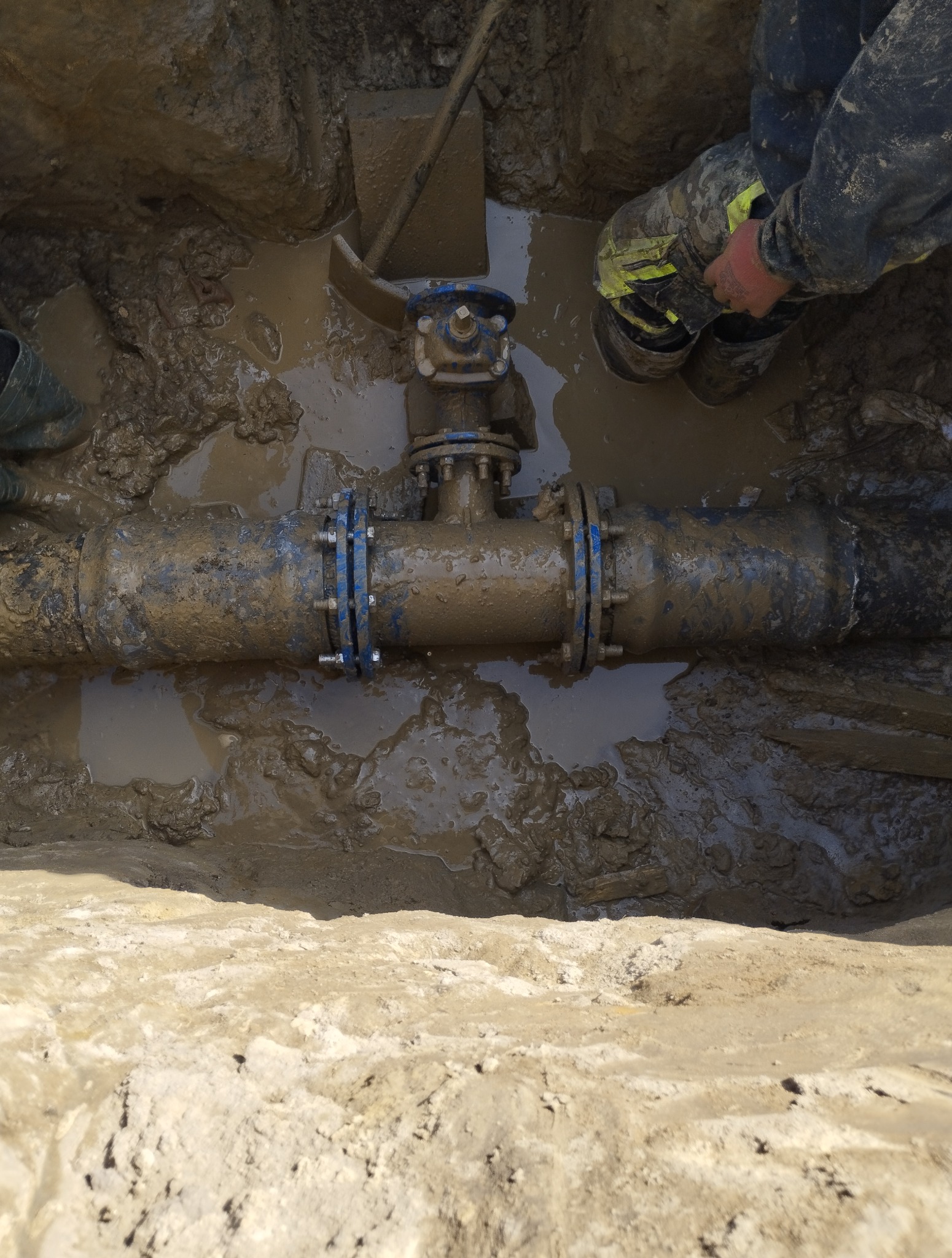 Rurociąg z zaworem w wykopie wypełnionym błotnistą wodą. Widoczne fragmenty ubrań i narzędzi hydraulika. Prace ziemne i instalacyjne w toku.