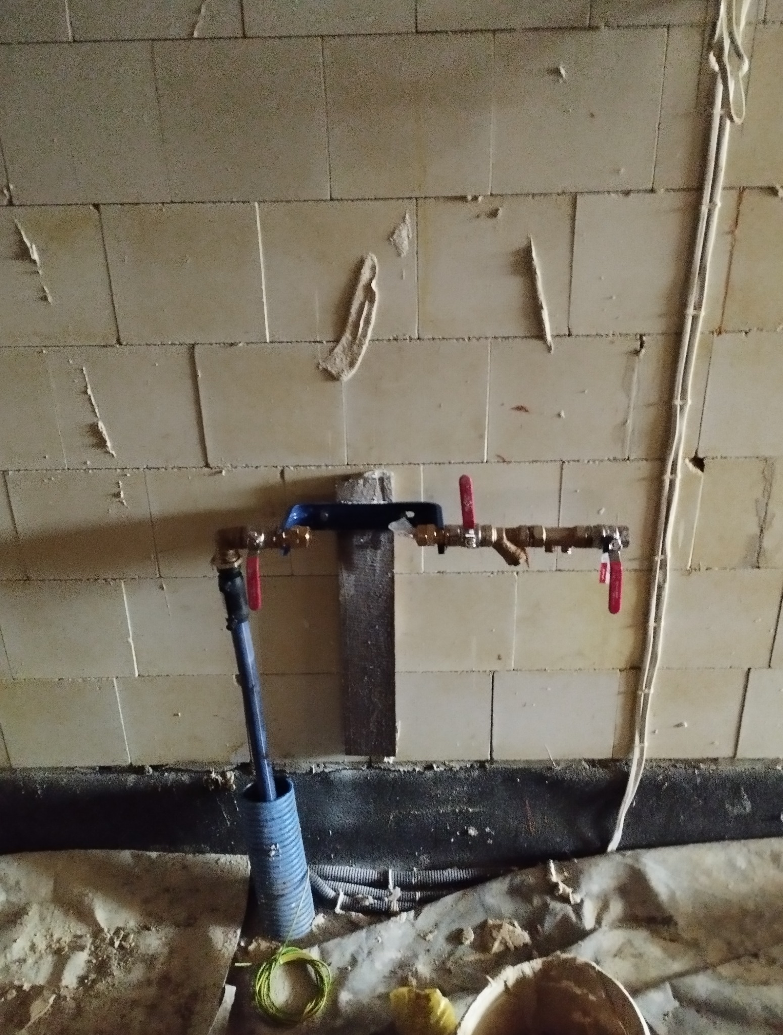 Instalacja wodna z zaworami odcinającymi na tle ściany z pustaków. Widoczne rury, zawory z czerwonymi uchwytami i niebieska rura osłonowa. Prace hydrauliczne w trakcie realizacji.