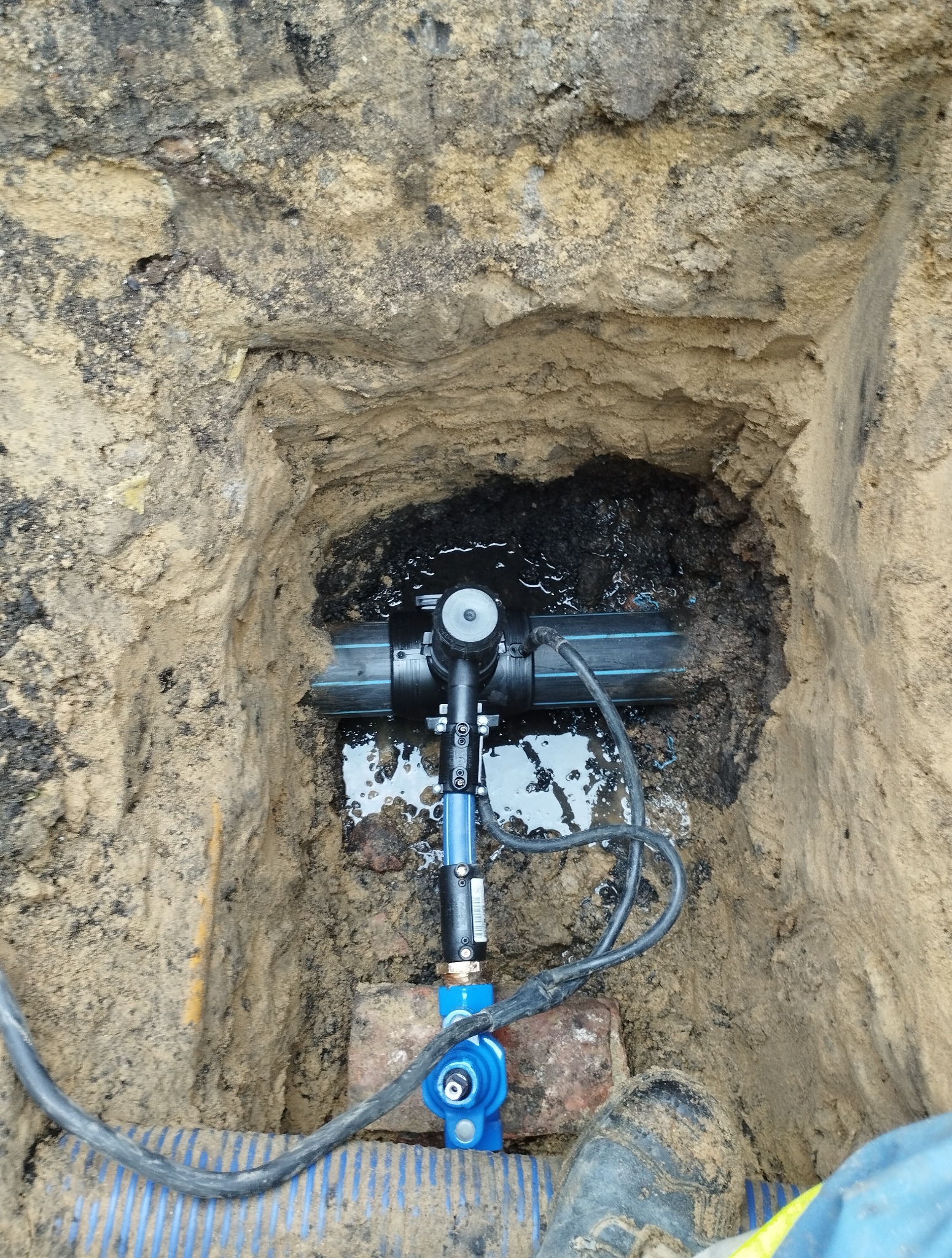 Widok z góry na nowo zamontowane, niebieskie zawory i czarne rury w wykopie z ziemi, częściowo wypełnionym wodą. Widoczny fragment buta roboczego.