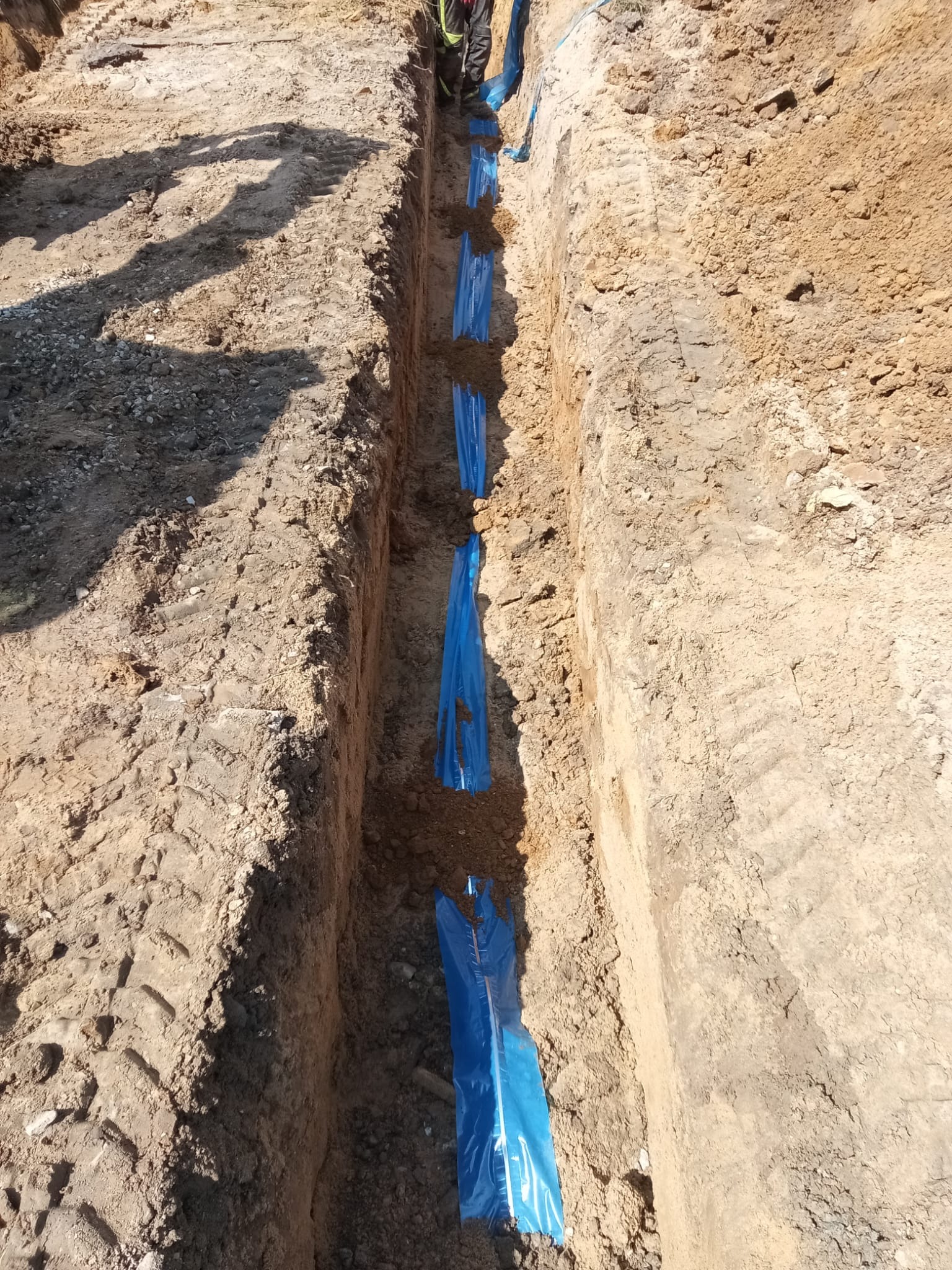 Wykop w ziemi z niebieską folią zabezpieczającą, prawdopodobnie przygotowany pod instalację rur. Widoczne ślady opon na ziemi i fragmenty sylwetek osób w tle.