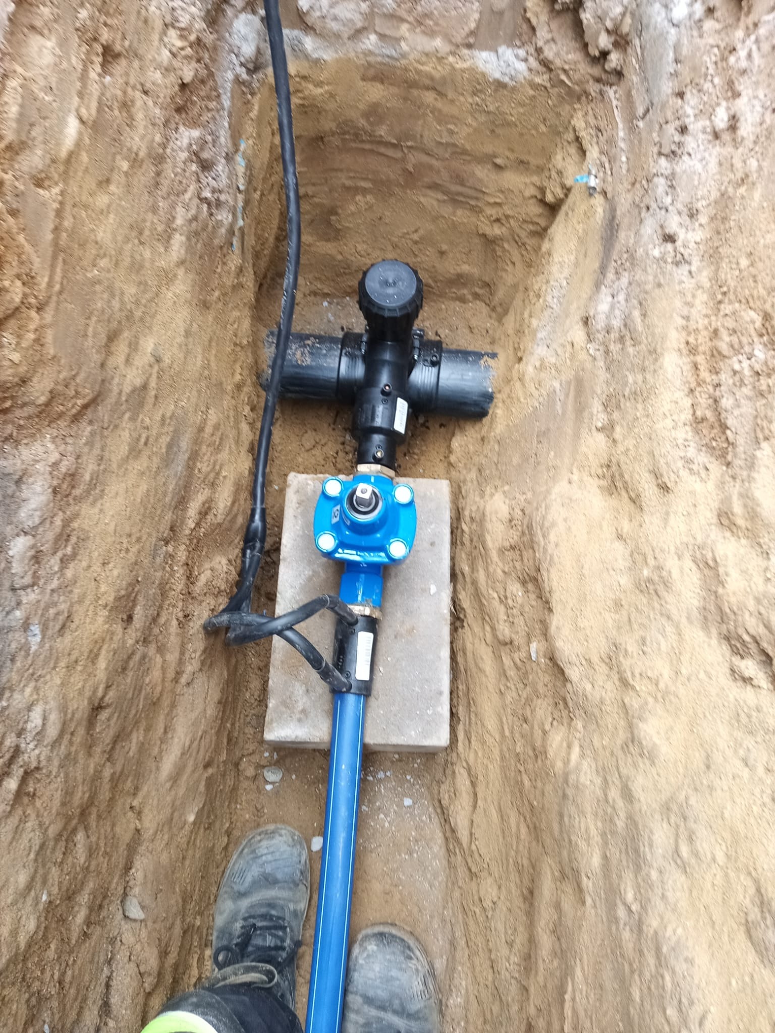 Niebieska rura wodociągowa z zaworem i czarnym przyłączem w wykopie ziemnym, osadzona na betonowej płycie. Widoczne buty osoby nadzorującej.