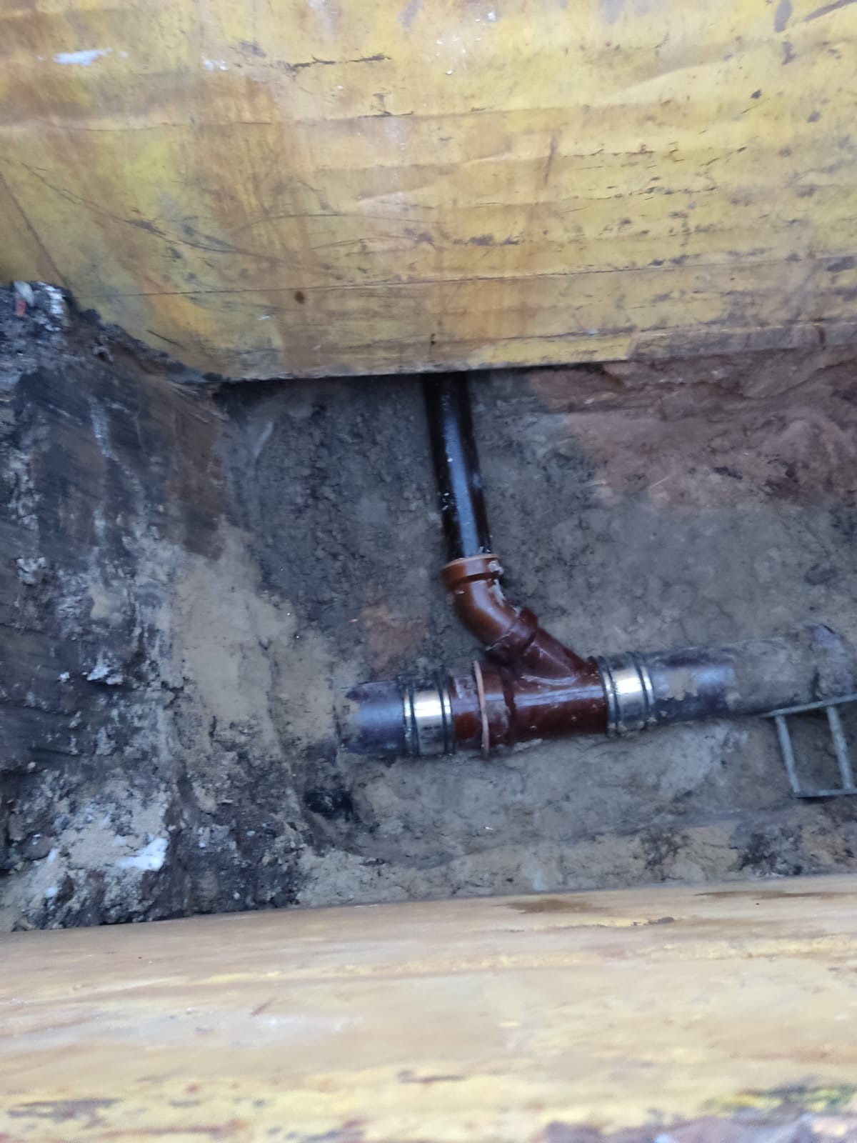 Brązowe rury kanalizacyjne z metalowymi złączkami ułożone w wykopie ziemnym, widoczne z góry. Fragment żółtej deski w tle.