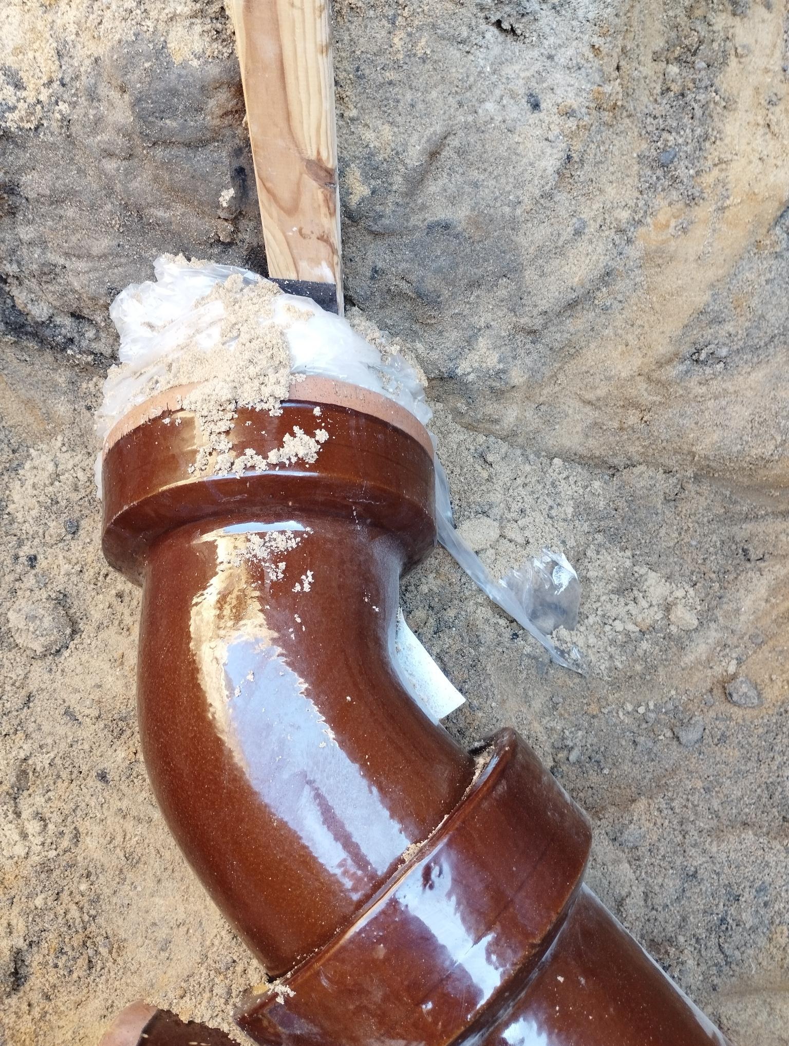 Brązowa, ceramiczna rura kanalizacyjna w wykopie, zabezpieczona folią i drewnianym stemplem. Widoczny piasek i ziemia. Element instalacji kanalizacyjnej.