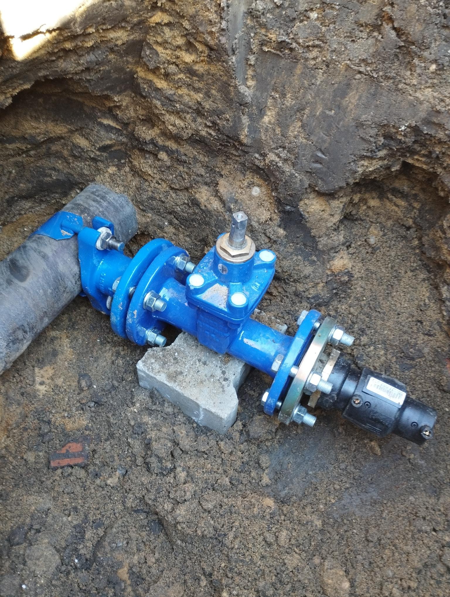 Niebieski zawór z rurą w wykopie, połączony śrubami. Widoczny fragment cegły i ziemia. Instalacja wodno-kanalizacyjna w trakcie montażu.