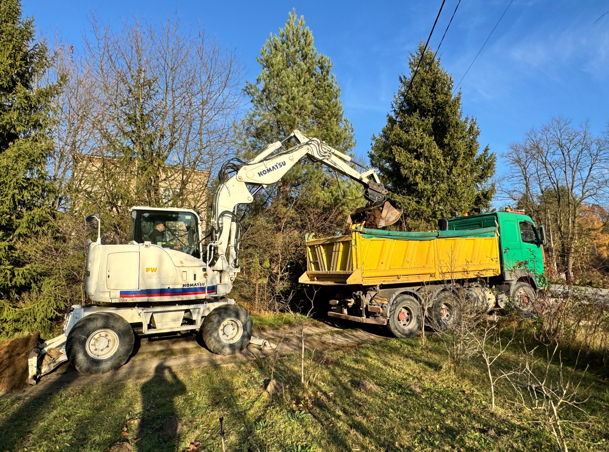 Biała koparka Komatsu ładuje ziemię do żółto-zielonej ciężarówki na tle drzew i błękitnego nieba. Widoczne szczegóły maszyny i otoczenia.