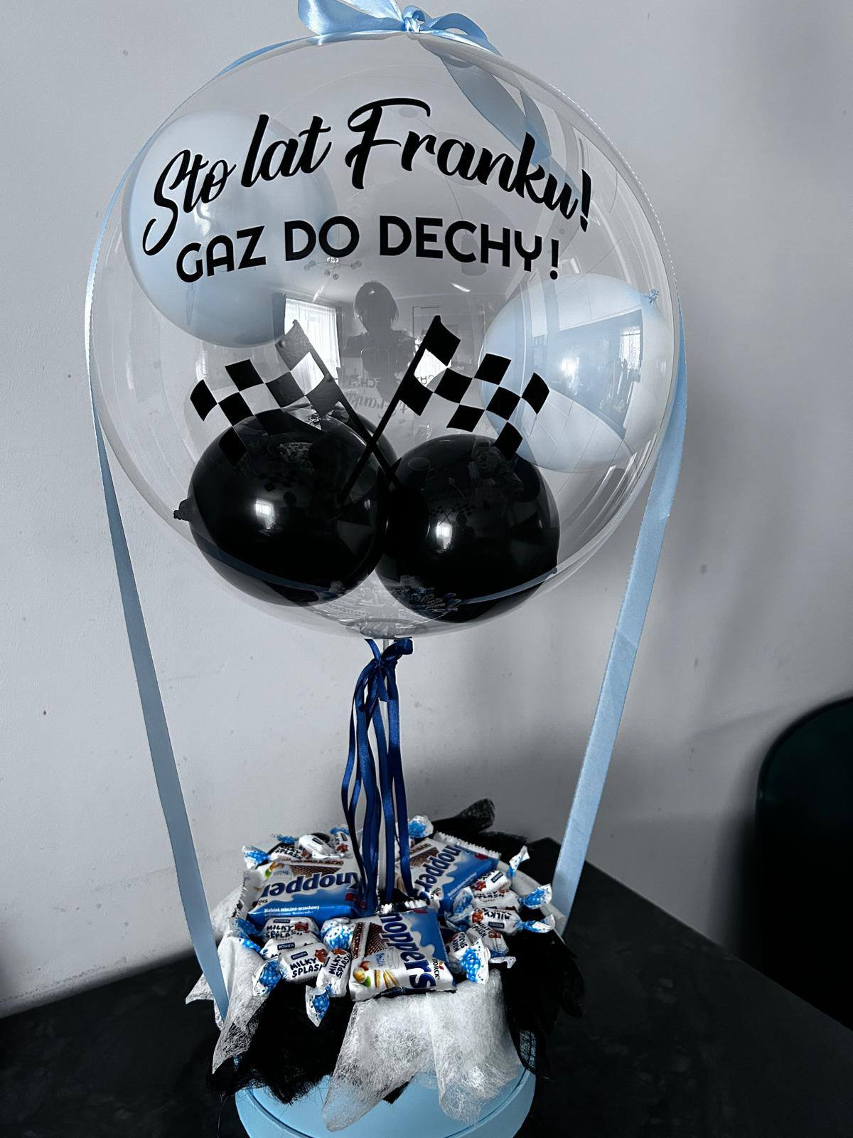 Ozdobny balon z napisem 'Sto lat Franku! GAZ DO DECHY!', z balonami w szachownicę i słodyczami w niebieskim pudełku, przewiązany błękitną wstążką.