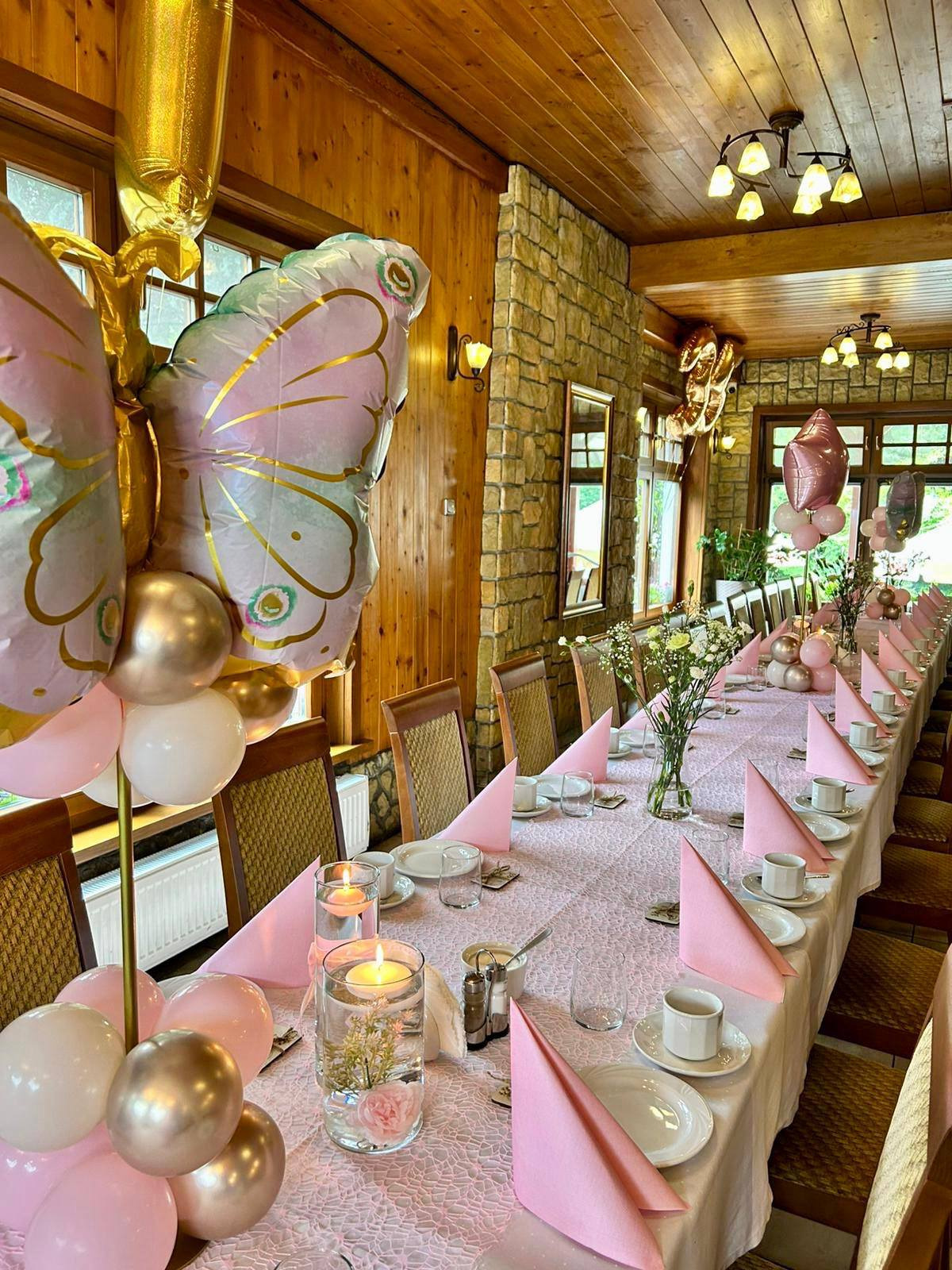Dekoracja stołu na imprezę w Czempiniu: różowe serwetki, balony w kształcie motyli, świece w szklanych naczyniach, kwiaty, zastawa stołowa w rustykalnym wnętrzu.