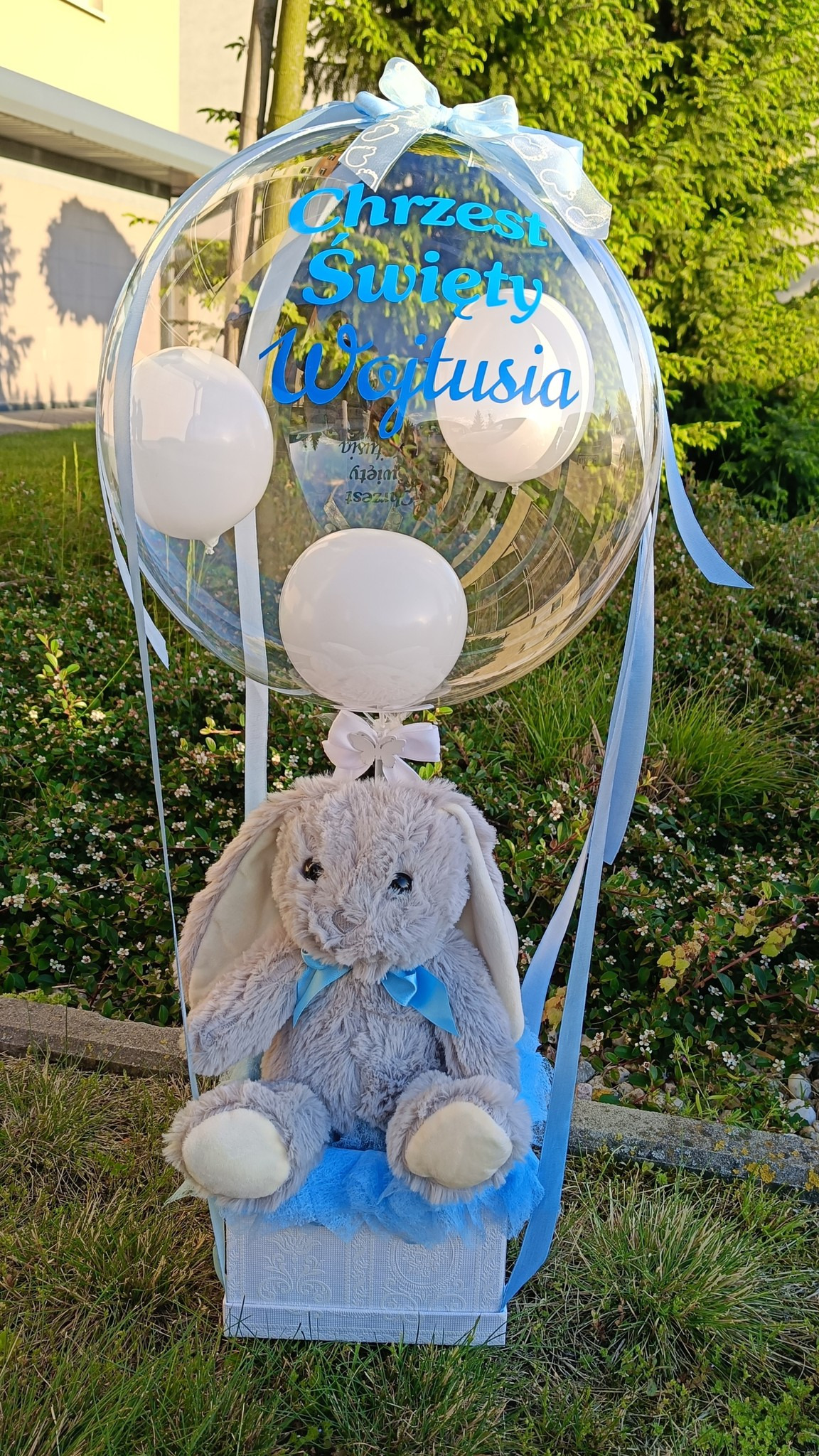 Dekoracja na chrzest: pluszowy królik w balonie z napisem 'Chrzest Święty Wojtusia', wstążki i białe balony. Całość w błękitnej tonacji, na tle zieleni.