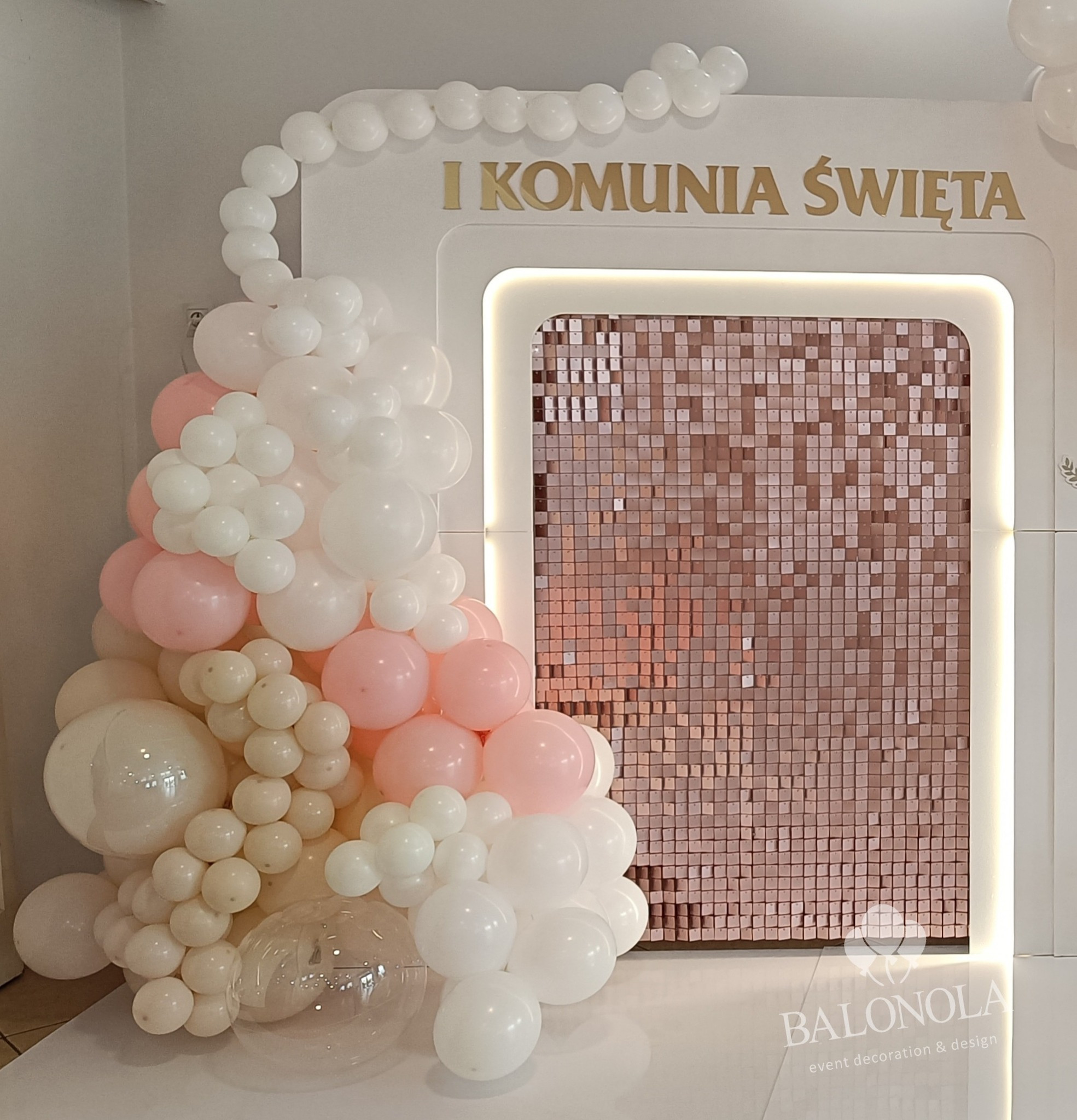 Dekoracja balonowa w bieli i różu, z napisem 'I Komunia Święta' na błyszczącym tle. Elegancka aranżacja na uroczystość.
