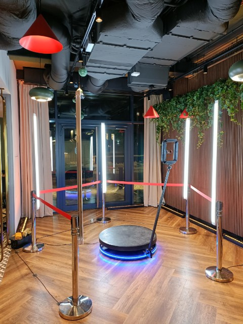 Okrągła platforma 360° z niebieskim podświetleniem, otoczona słupkami z czerwonymi taśmami, gotowa do zabawy podczas imprezy w nowoczesnym wnętrzu.