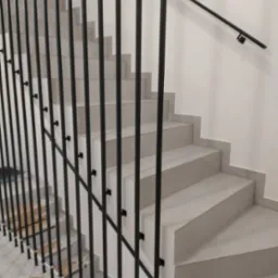 Nowoczesne schody z szarymi stopniami i czarną, metalową balustradą z pionowych prętów. Minimalistyczny design, jasne tło ściany.