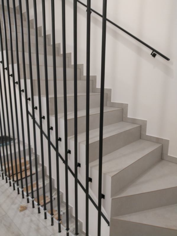 Nowoczesne schody z szarymi stopniami i czarną, metalową balustradą z pionowych prętów. Minimalistyczny design, jasne tło ściany.