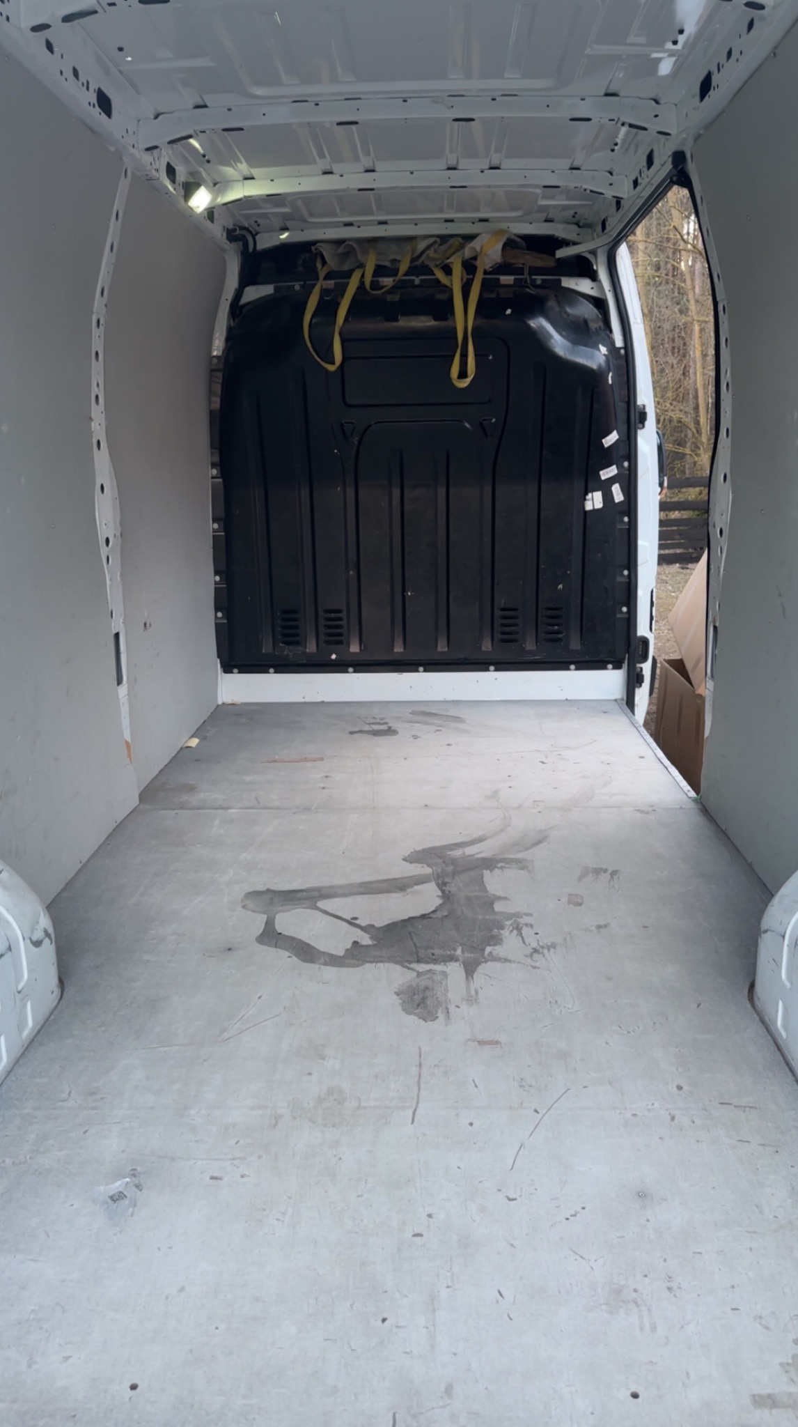 Wnętrze paki dostawczego busa z widocznymi pasami zabezpieczającymi ładunek i plamami na podłodze. Otwarta przestrzeń ładunkowa z widokiem na zewnątrz.