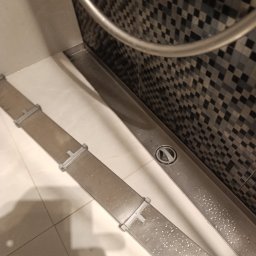 Effect cleaning - Umyta, stalowa listwa odpływowa prysznica z widocznymi kroplami wody, na tle mozaiki z szarych kwadratów. Ujęcie z góry, pod kątem.