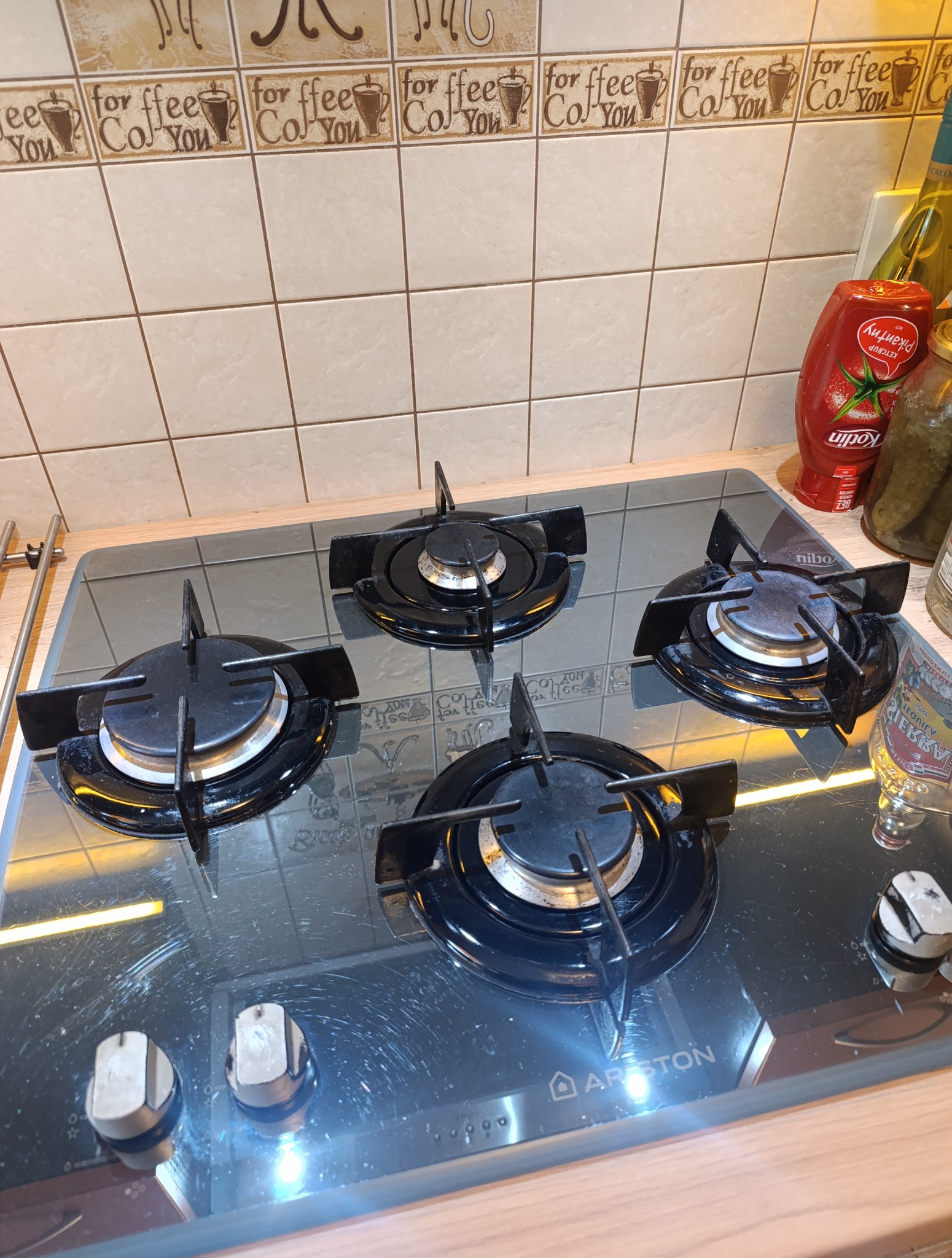 Czyszczenie płyty gazowej Ariston z czterema palnikami, na tle płytek z motywem kawy. Widoczne ślady po czyszczeniu.