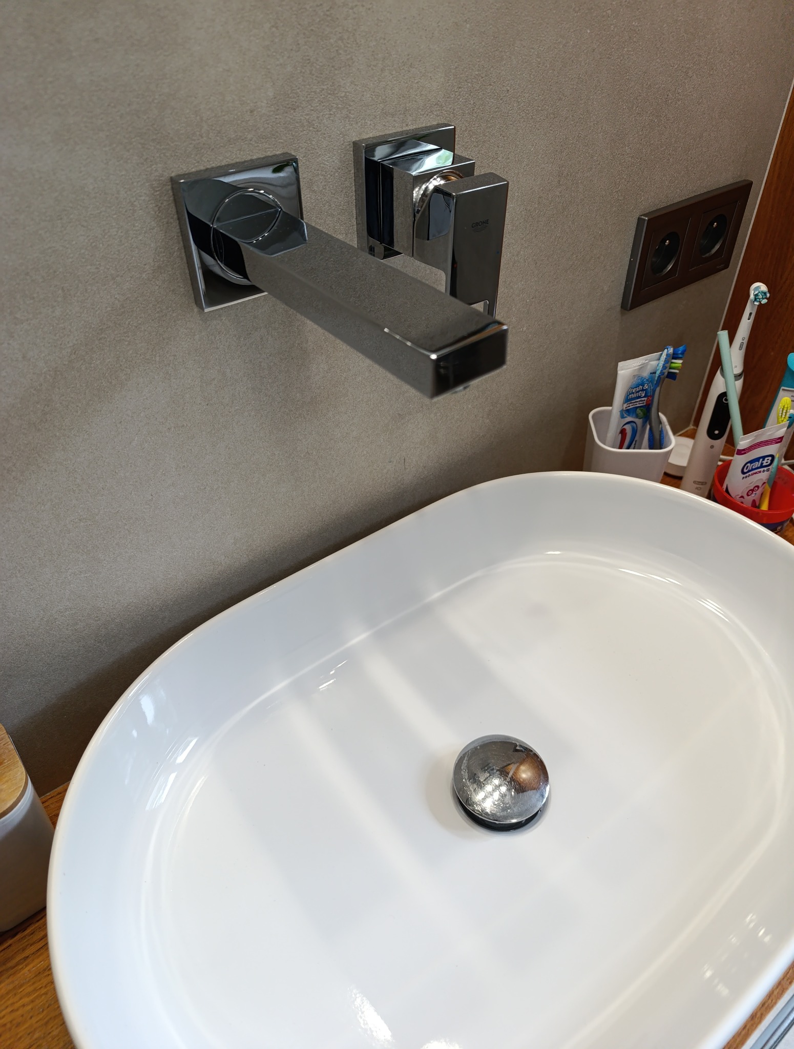 Biała, owalna umywalka z chromowaną baterią Grohe na beżowej ścianie. Widoczne gniazdko elektryczne i przybory toaletowe. Minimalistyczny design łazienki.