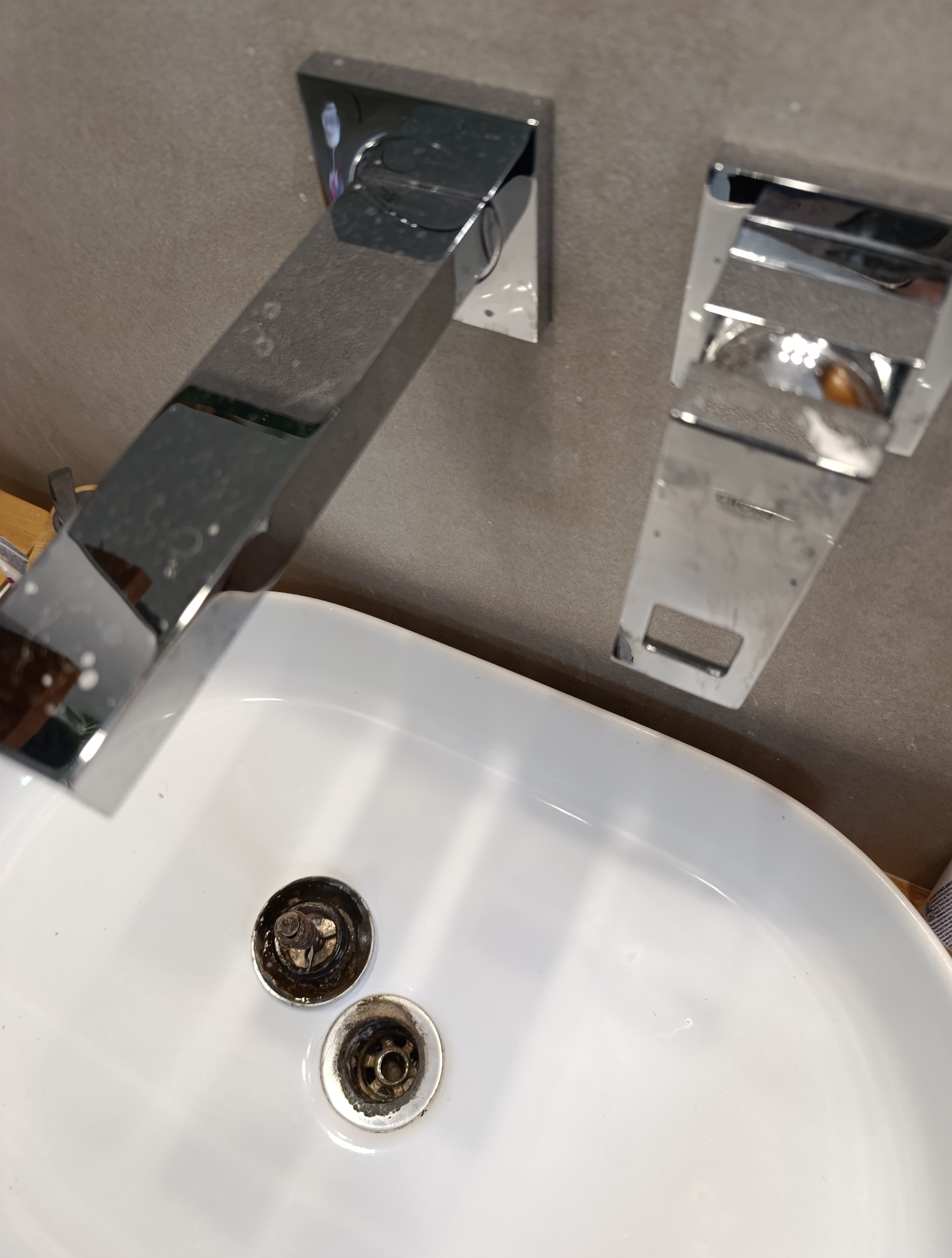 Brudna, biała umywalka z chromowaną armaturą i zatkanymi odpływami przed czyszczeniem. Widoczne zacieki i osad.