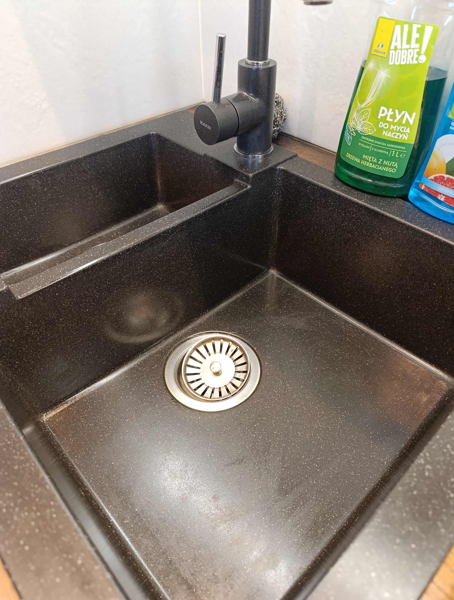 Czarny, dwukomorowy zlew granitowy z baterią Blancco i płynem do naczyń o zapachu mięty i drzewa herbacianego. Widoczny odpływ i fragment blatu.