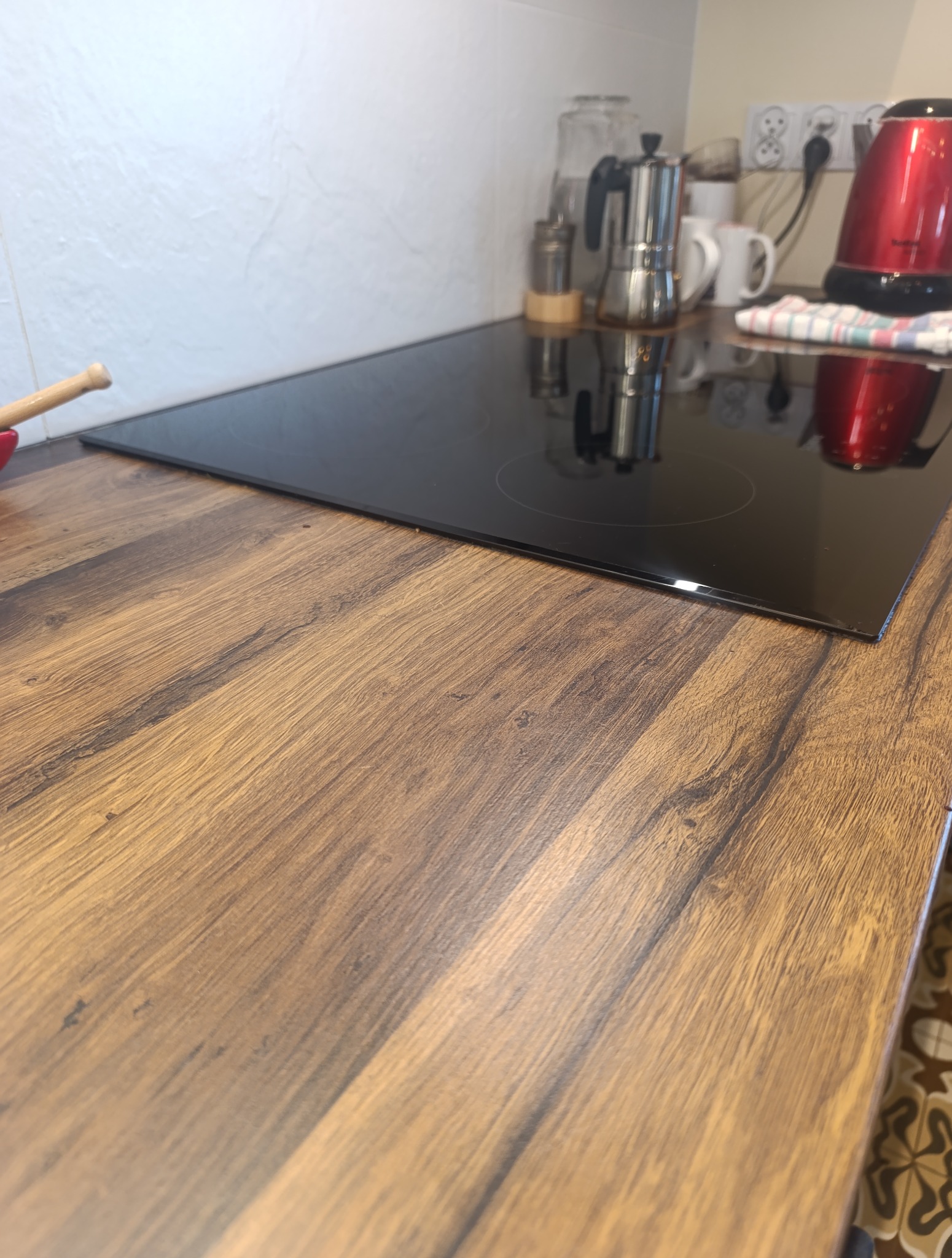 Błyszcząca płyta indukcyjna na drewnianym blacie kuchennym z widocznymi akcesoriami: czajnik, kawiarka i naczynia. Czysta powierzchnia gotowa do użycia.