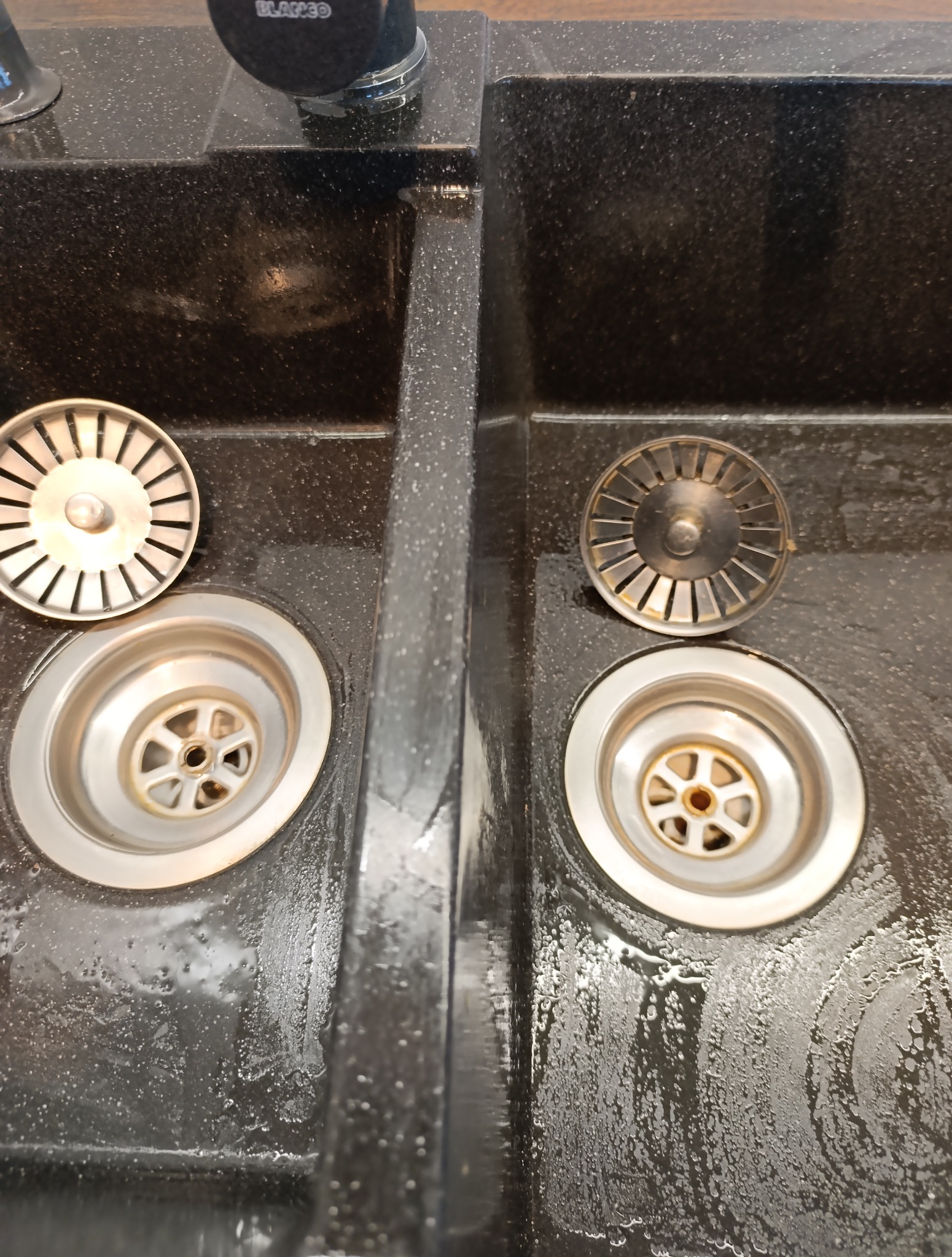 Błyszczący, czarny, dwukomorowy zlewozmywak granitowy z metalowymi odpływami, widoczne krople wody po czyszczeniu. Detal po sprzątaniu.