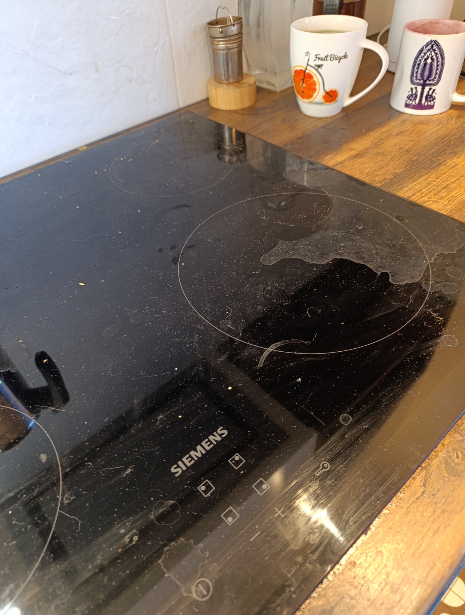 Zabrudzona płyta indukcyjna marki Siemens z widocznymi plamami, smugami i drobnymi zabrudzeniami na czarnej powierzchni. W tle kubki i akcesoria kuchenne.