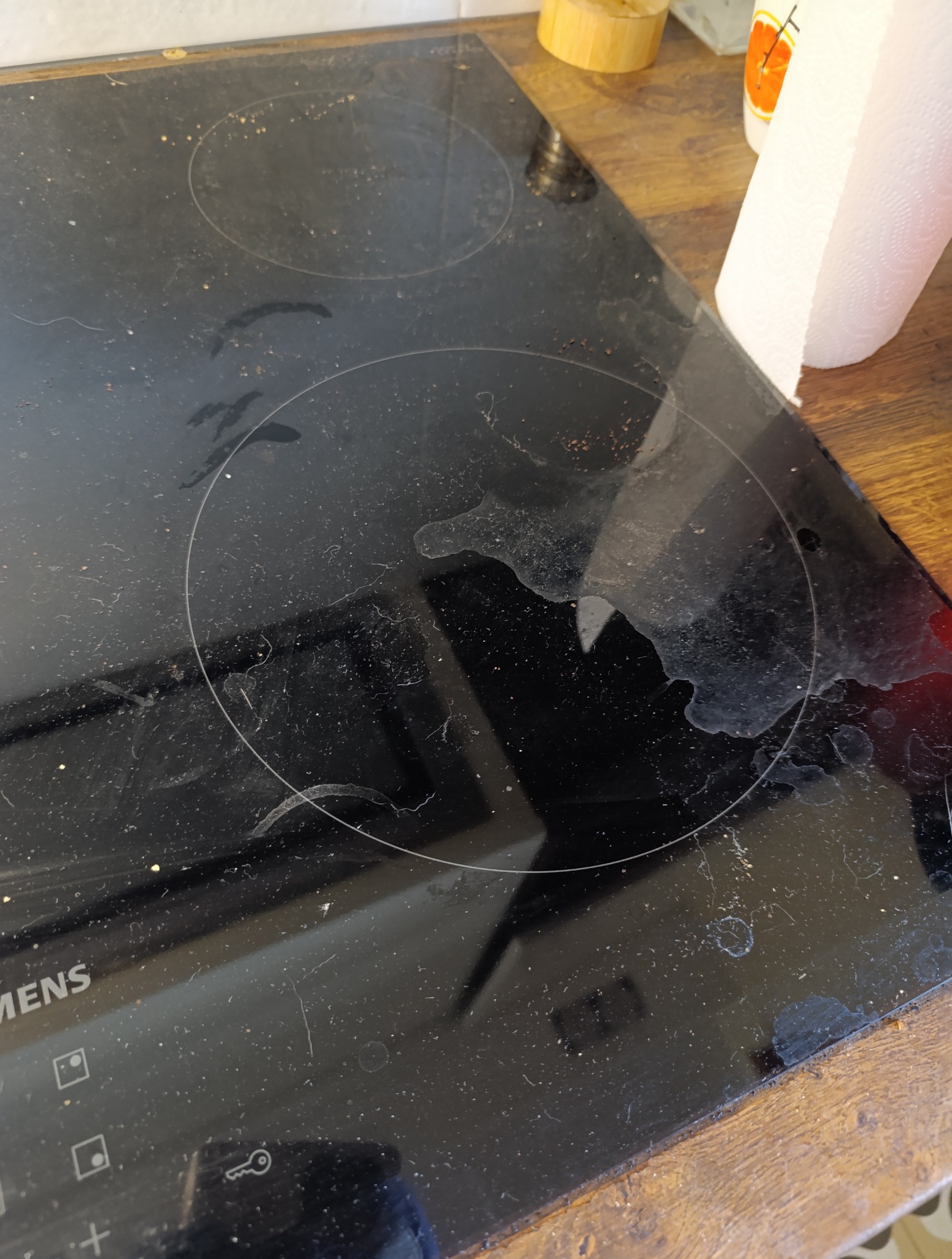 Zabrudzona płyta indukcyjna Siemens z resztkami jedzenia i smugami, widoczny fragment blatu i ręcznik papierowy. Niezadbana kuchnia przed czyszczeniem.