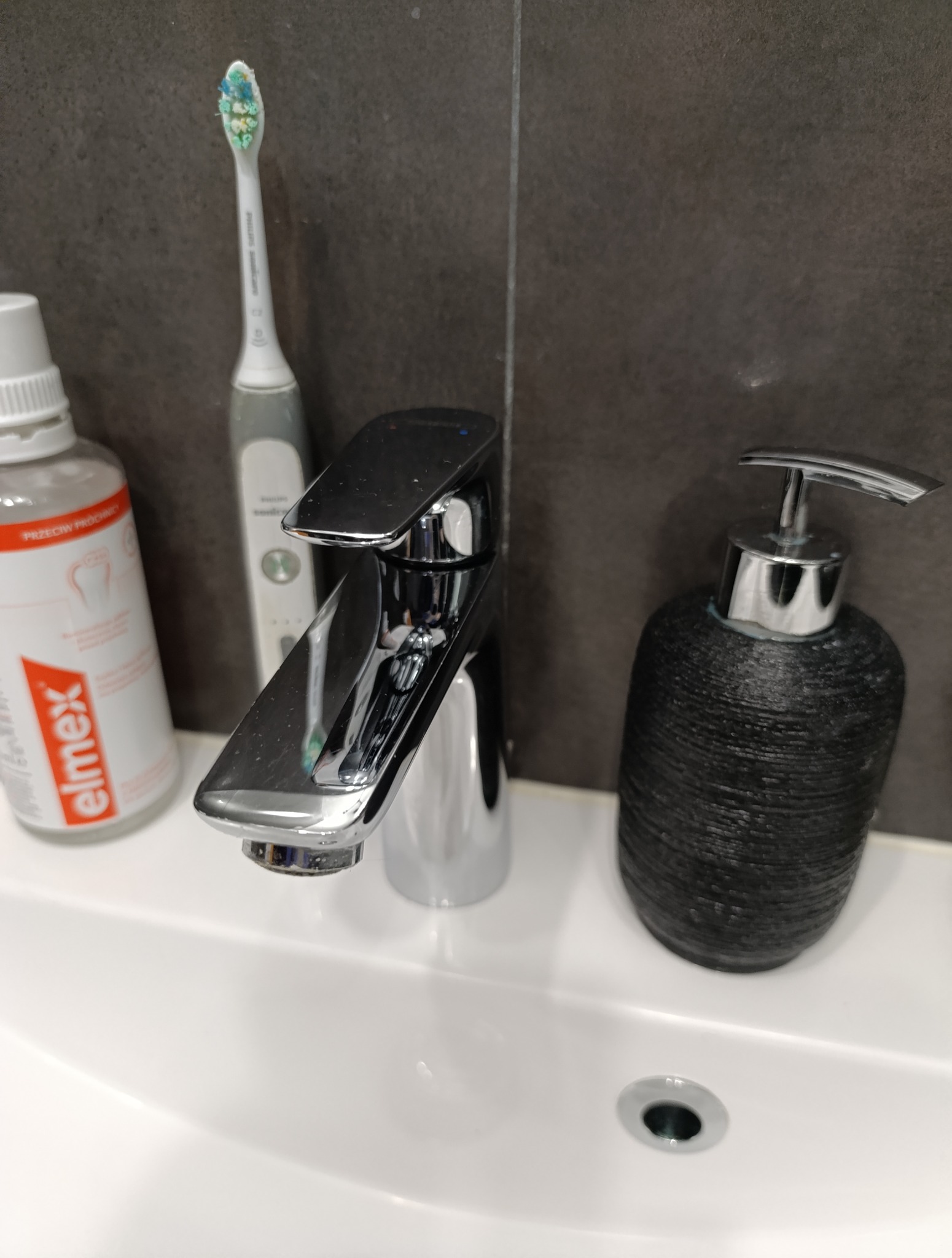 Bateria umywalkowa, obok szczoteczka elektryczna, płyn do płukania ust Elmex i dozownik mydła na tle szarych płytek. Widok na czystą umywalkę.