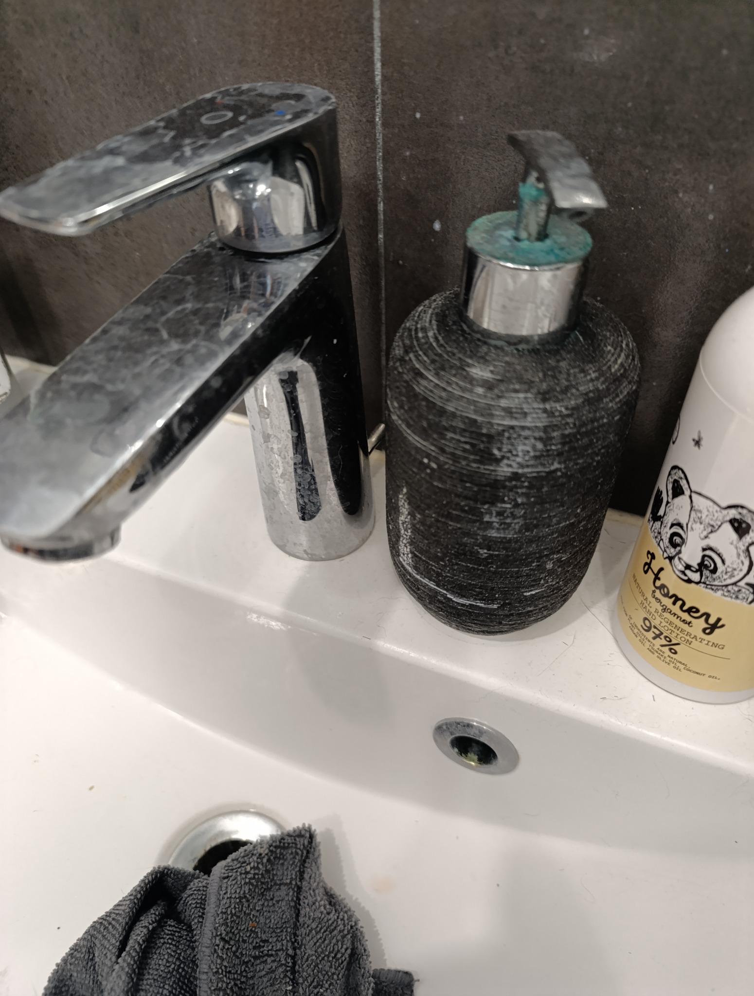 Z bliska: zacieki na chromowanej baterii umywalkowej, dozowniku mydła i resztki włosów w umywalce. Widok przed czyszczeniem.