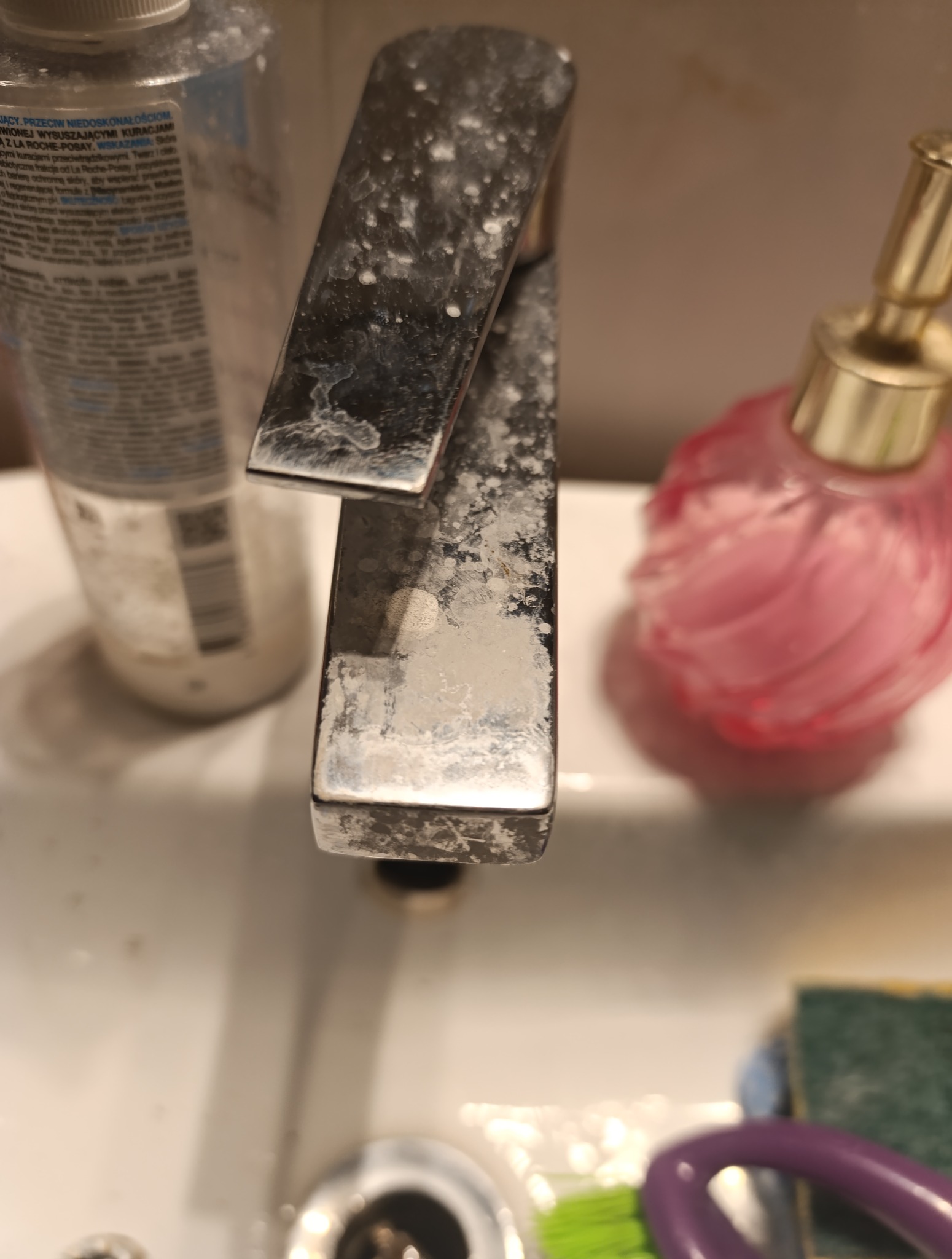 Zakamieniona bateria umywalkowa z bliska, przed czyszczeniem. Widoczne osady i zacieki. Obok butelka kosmetyku i gąbka do czyszczenia. Słabe oświetlenie.