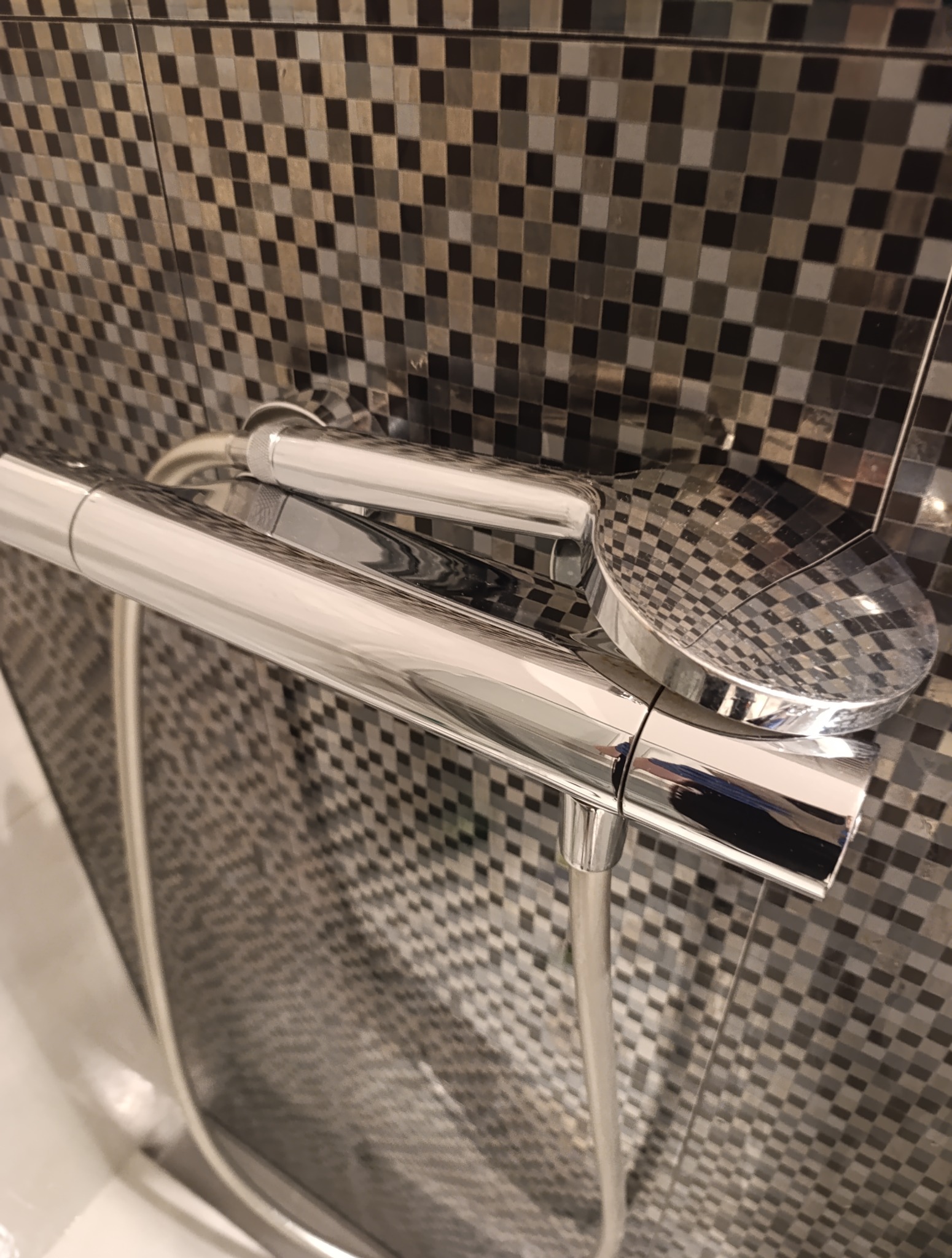 Błyszcząca, chromowana bateria prysznicowa z deszczownicą na tle mozaiki w odcieniach szarości i czerni. Widoczna słuchawka i wąż prysznicowy.