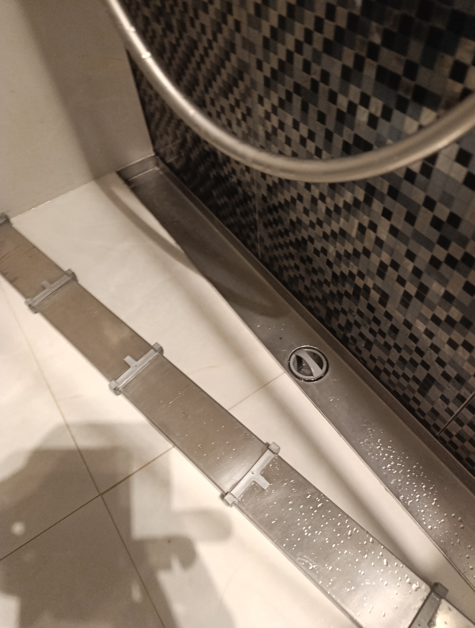 Umyta, stalowa listwa odpływowa prysznica z widocznymi kroplami wody, na tle mozaiki z szarych kwadratów. Ujęcie z góry, pod kątem.