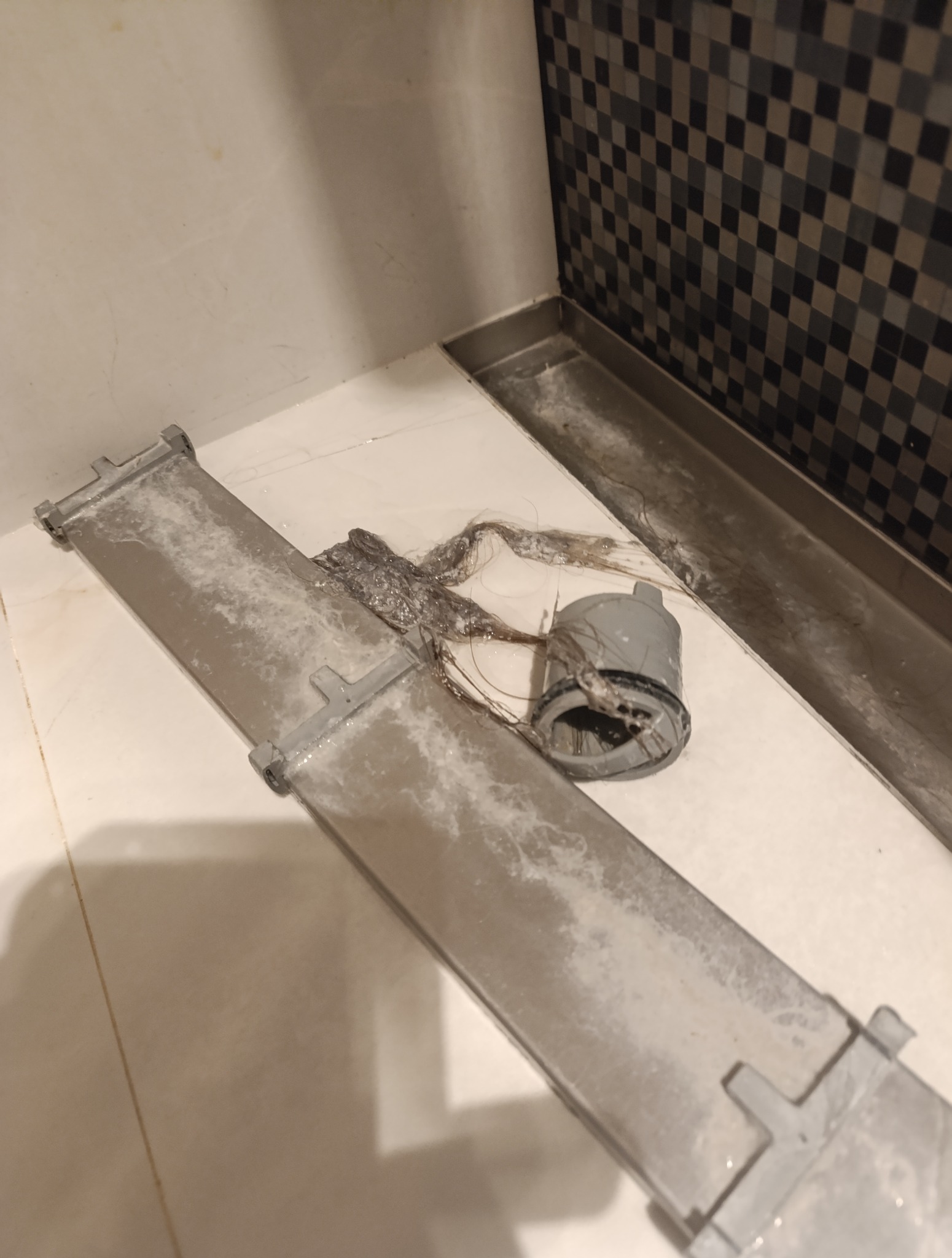 Udrożniony odpływ liniowy prysznica z wyjętym syfonem i zebranymi włosami. Czyszczenie odpływu w łazience z mozaiką.