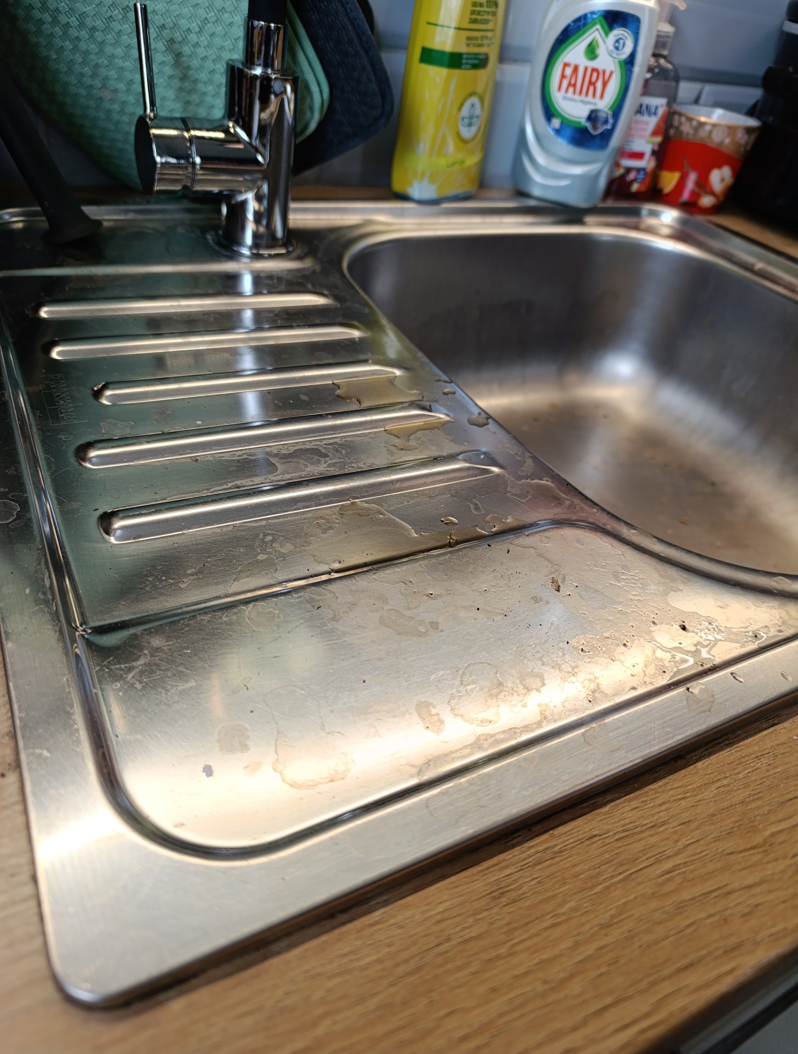 Zlewozmywak stalowy przed czyszczeniem. Widoczne zaschnięte plamy i zabrudzenia na powierzchni. Kran w tle. Ujęcie z bliska, ukazujące detale.