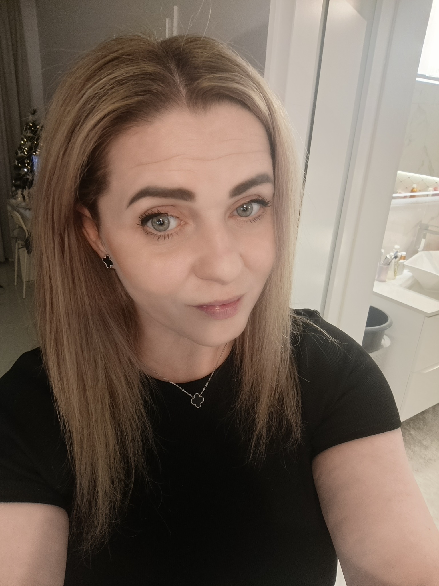 Portret kobiety z blond włosami, w czarnej bluzce, z subtelną biżuterią, na tle łazienki z elementami dekoracyjnymi i choinką w tle. Ujęcie z bliska, naturalne światło.