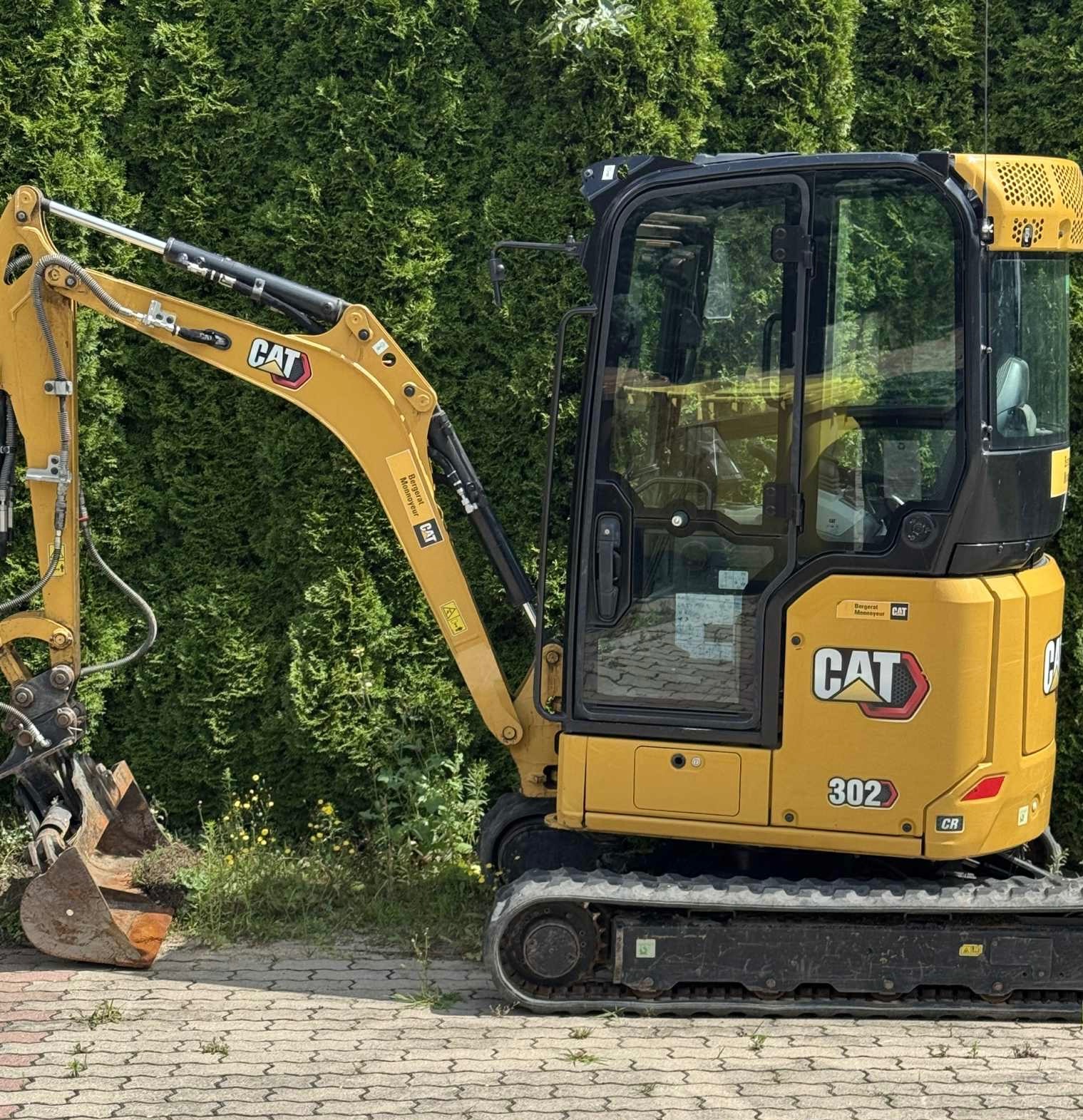 Żółta minikoparka CAT 302 CR na kostce brukowej, na tle zielonego żywopłotu. Widok z boku, lekko od przodu. Widoczny lemiesz i kabina operatora.