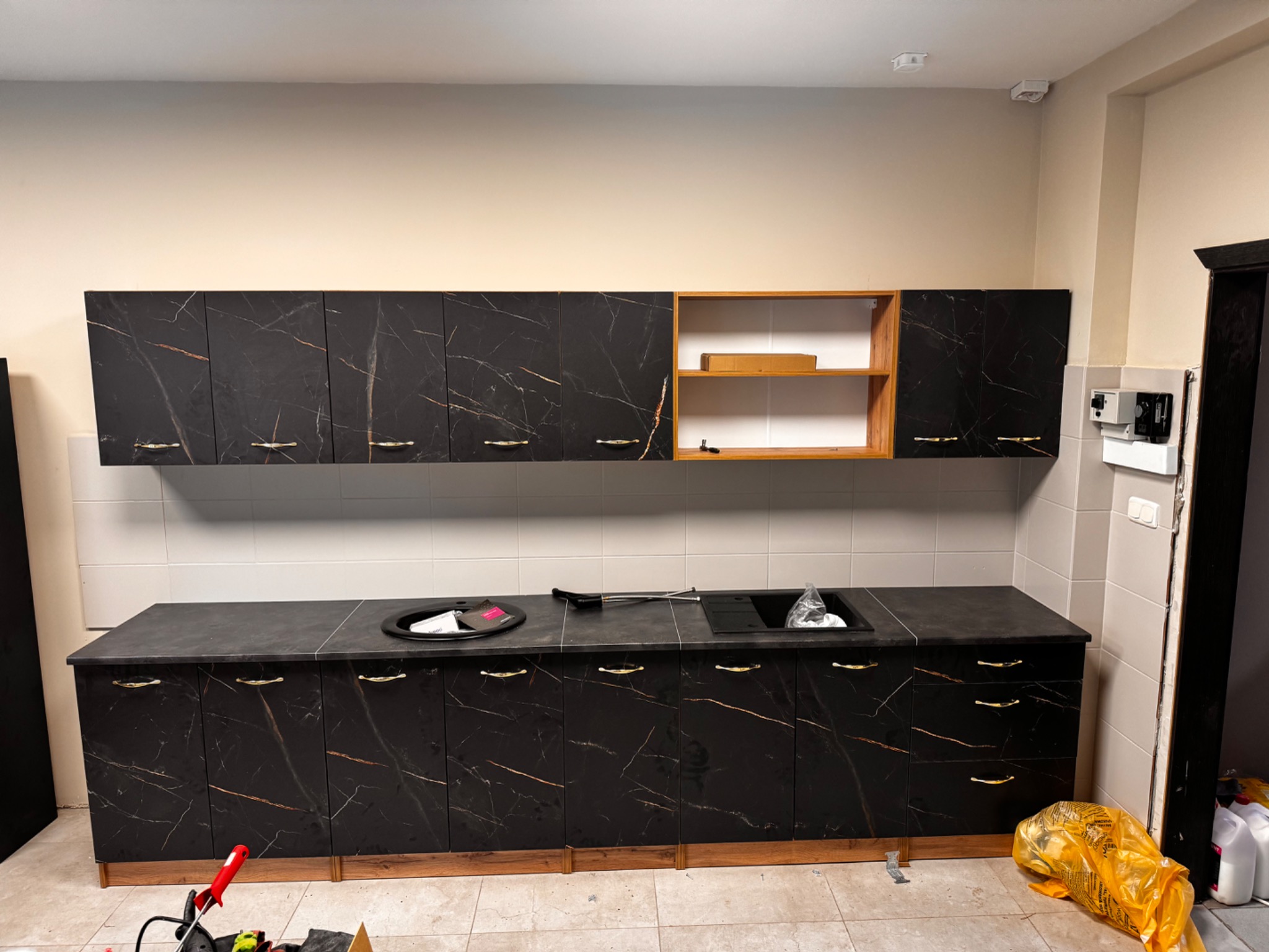 Czarne szafki kuchenne z marmurowym wzorem i złotymi uchwytami, zamontowane na tle białych płytek. Częściowo widoczne narzędzia i materiały budowlane.