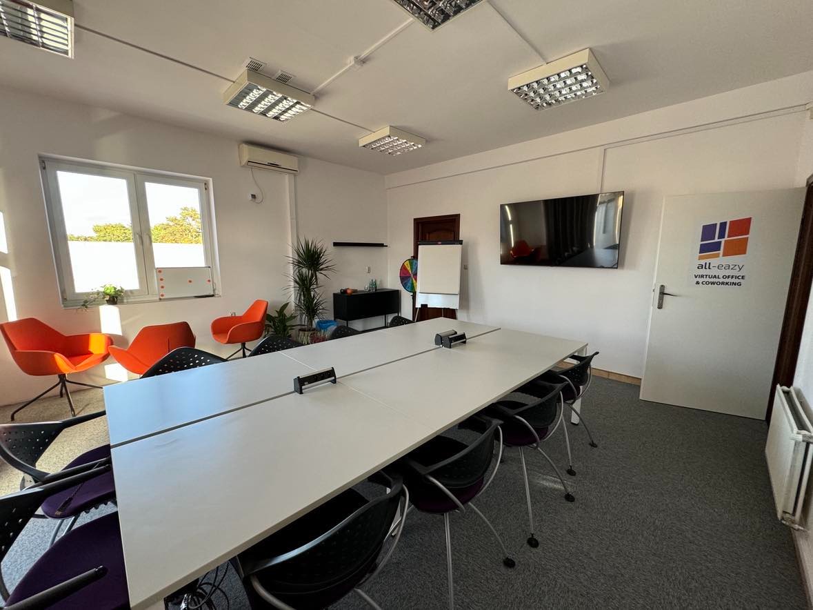 Jasne biuro coworkingowe z długim białym stołem, krzesłami, pomarańczowymi fotelami i logo firmy na drzwiach. Minimalistyczny design.