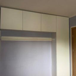 Niewczas Meble - Biała zabudowa meblowa z szafkami wiszącymi i otwartą wnęką z drążkiem na ubrania, na tle jasnofioletowej ściany i drewnianej podłogi w Józefowie.