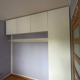 Niewczas Meble - Nowoczesna biała zabudowa szafy na wymiar z wiszącymi szafkami i drążkiem, w pokoju z jasnofioletowymi ścianami i drewnianą podłogą, obok drewnianych drzwi.