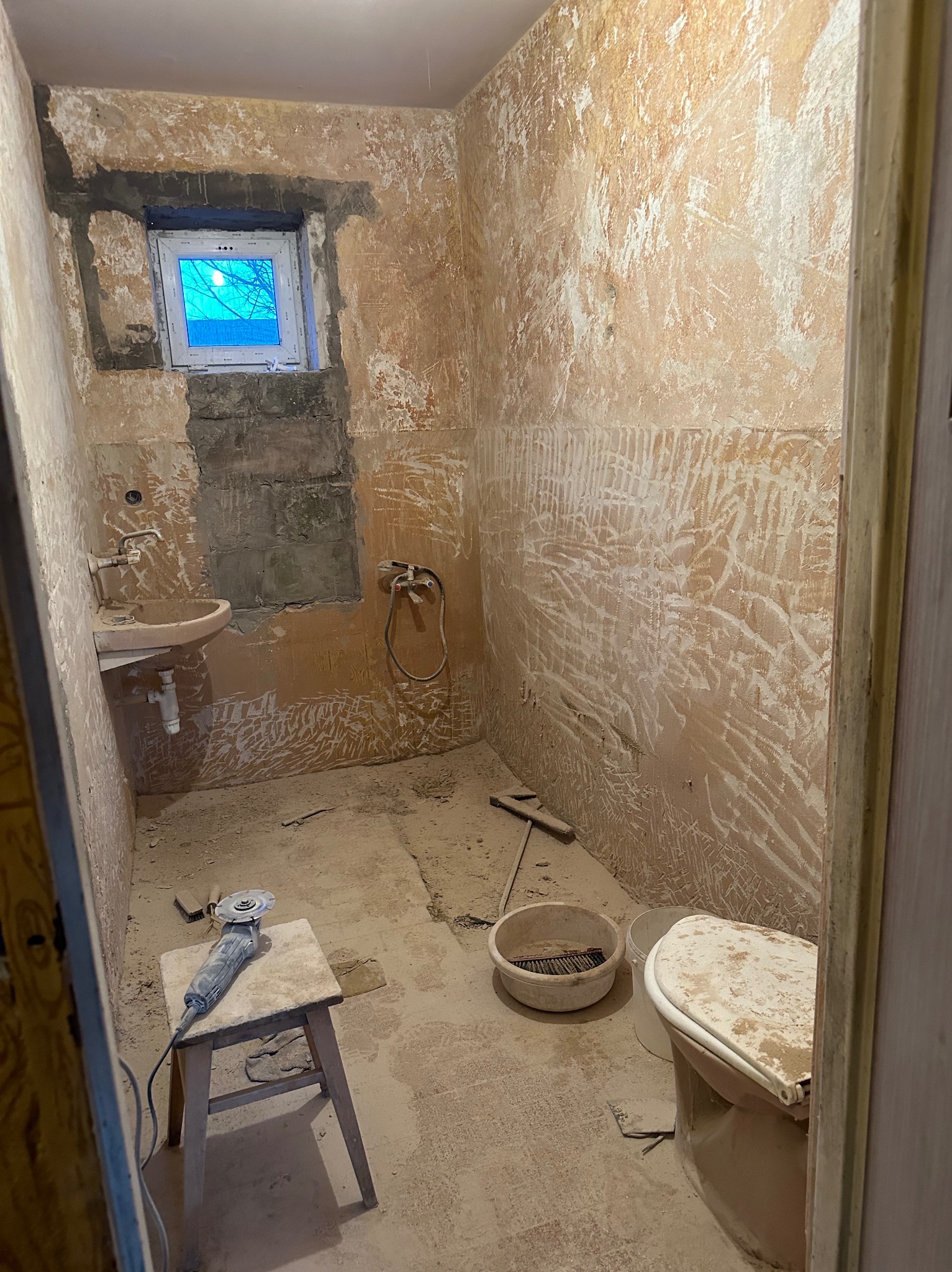 Łazienka w trakcie gruntownego remontu: usunięte powłoki malarskie, wymienione okno, przygotowane ściany pod gładzie. Widoczne narzędzia i stare sanitariaty.