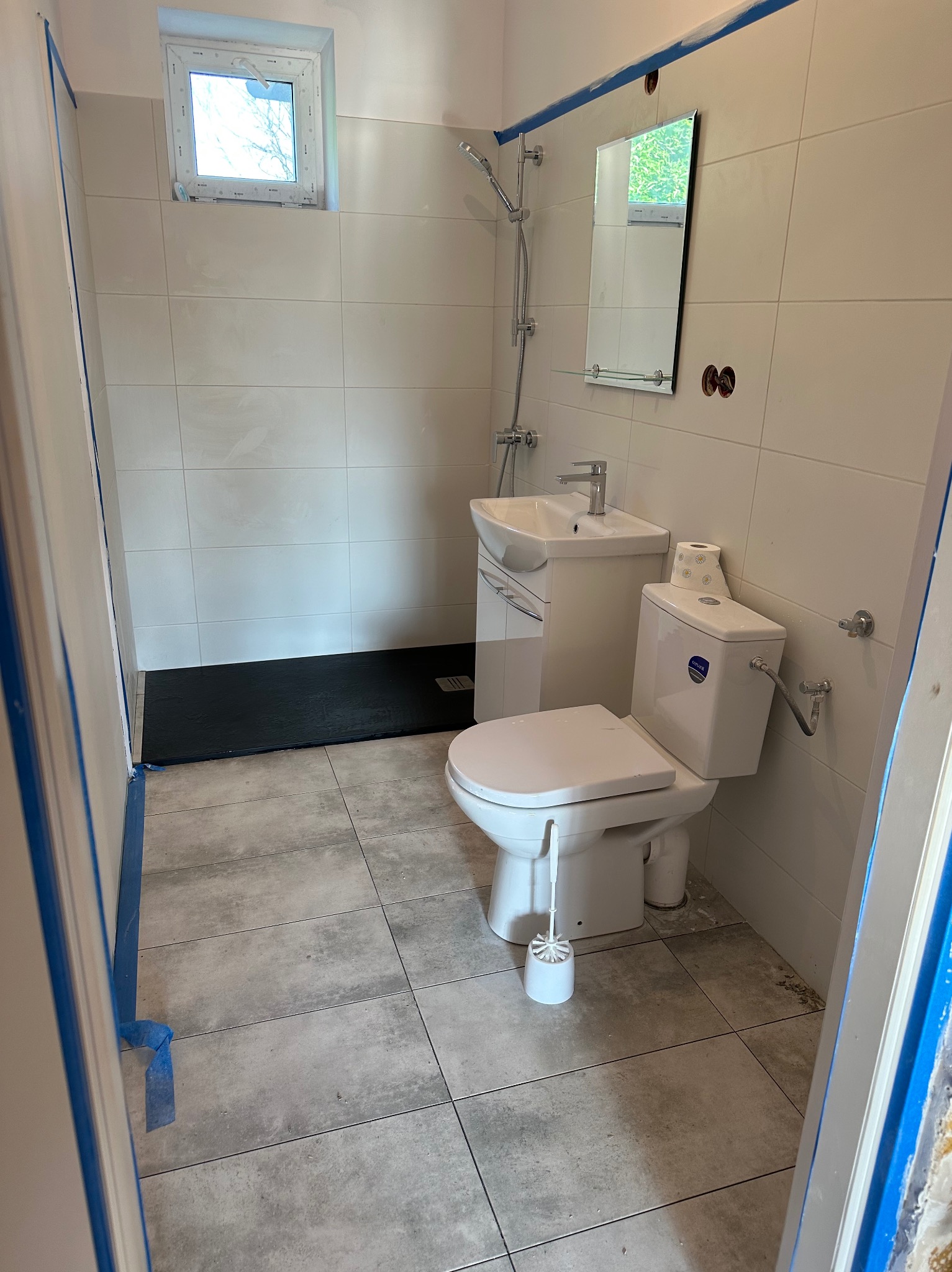 Wykończona łazienka z czarnym brodzikiem, umywalką z szafką i toaletą. Ściany wyłożone białymi płytkami. Otwór okienny i lustro na ścianie. Podłoga wyłożona szarą terakotą.