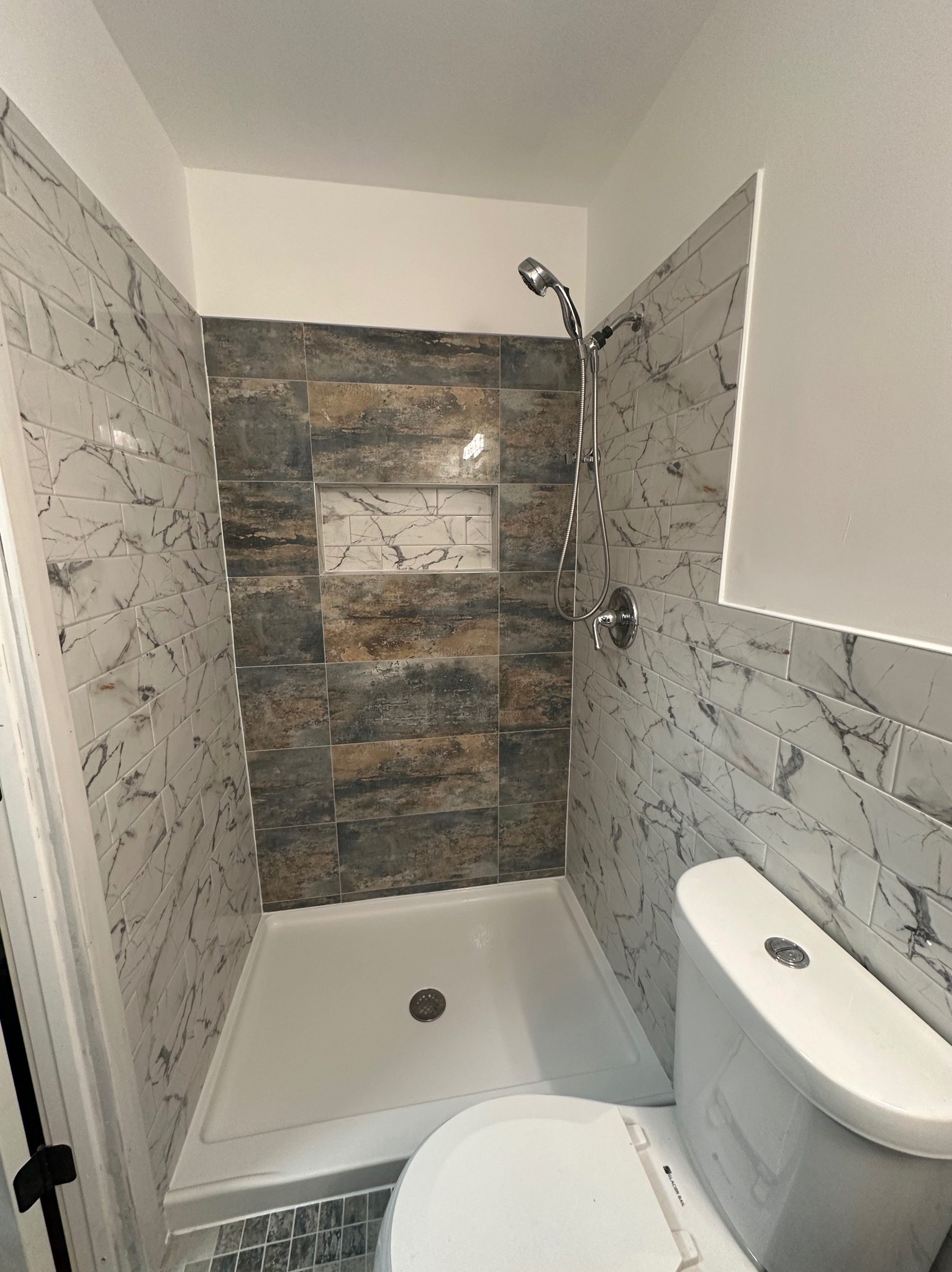 Wykończona kabina prysznicowa z marmurowymi płytkami i wnęką, widok z toaletą. Uwagę przykuwa mozaika podłogowa i chromowana armatura.