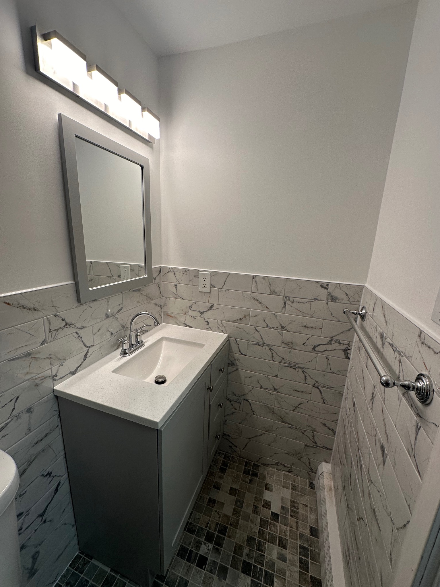 Wykończona łazienka z szarą szafką pod umywalką, marmurowymi płytkami na ścianach i mozaikową podłogą. Nad umywalką lustro z oświetleniem.