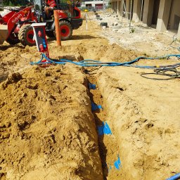 Elgrom - Wykop z niebieskimi rurami w ziemi, obok czerwony agregat prądotwórczy i koparka Kubota. Widoczne kable i przewody. Prace ziemne na budowie.