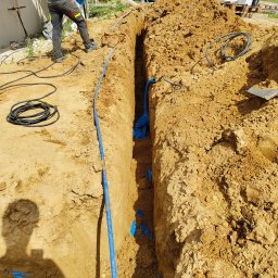 Elgrom - Wykop z ułożonymi niebieskimi rurami kablowymi i czarnym przewodem elektrycznym. Widoczny cień osoby i fragment sylwetki pracownika w kombinezonie.